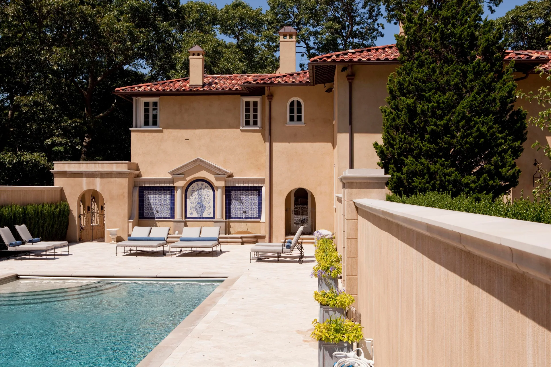 An outdoor swimming pool and stone patio with grey lounge chairs next to a beige stucco Mediterranean house with a blue tile wall mural.