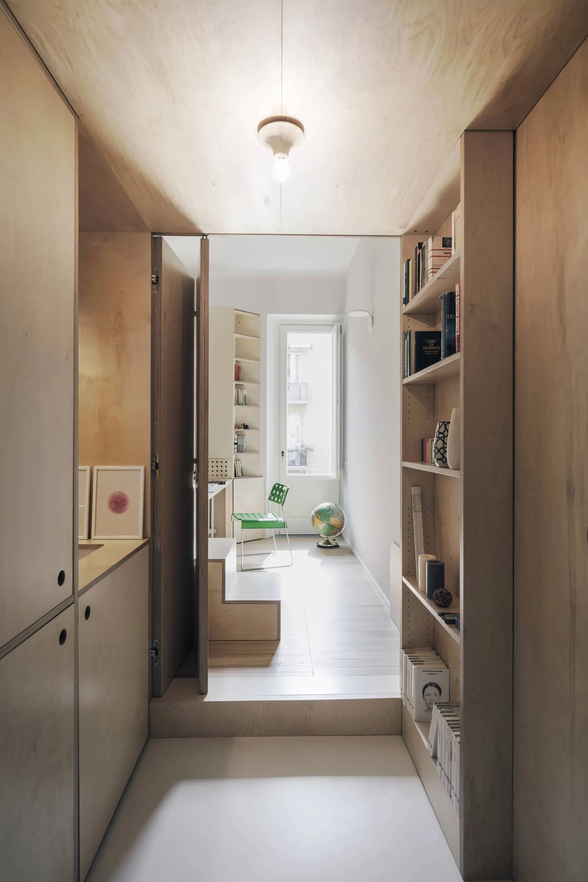 A narrow hallway featuring custom plywood wall paneling and built-in shelving leads into a bright white room with a green chair.