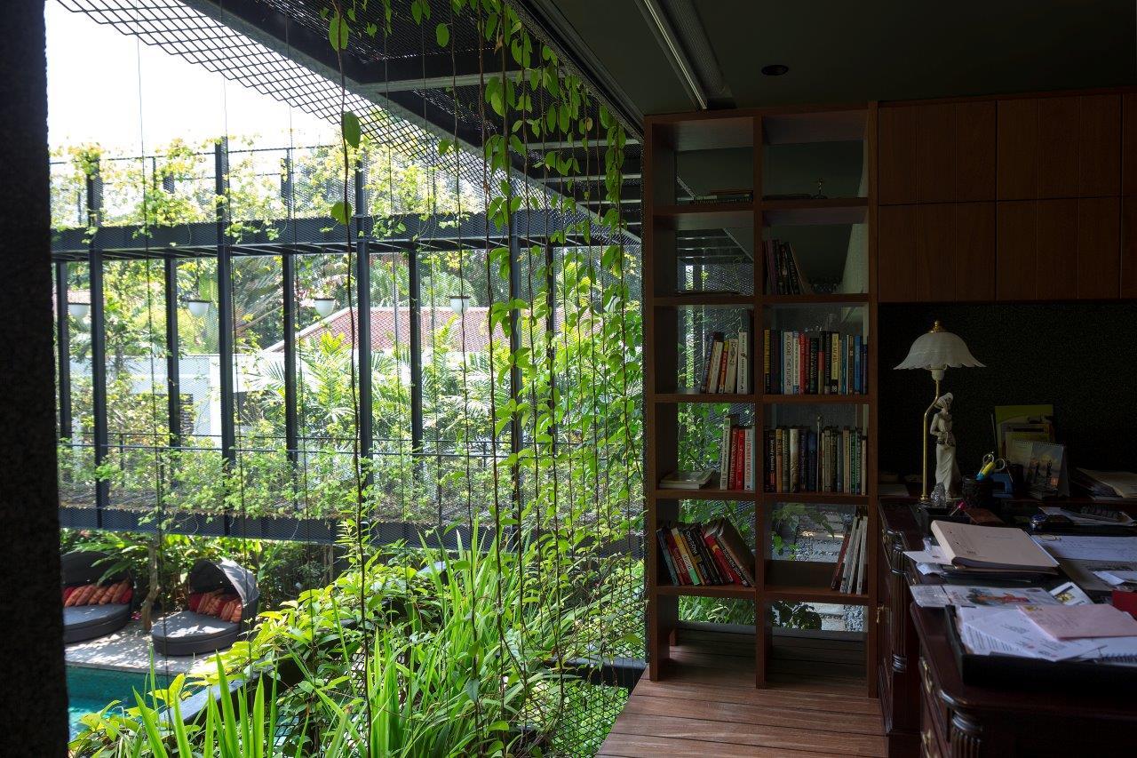 A home office features a dark wood desk and an open-backed wooden bookshelf next to a glass wall overlooking lush green vines and an outdoor pool.