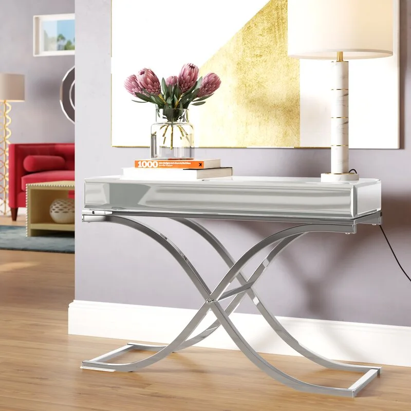 A mirrored console table with a polished chrome x-frame base stands against a lavender wall decorated with gold abstract art and a white lamp.