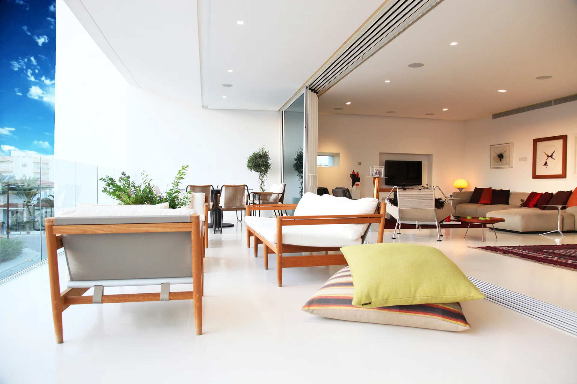 A modern open-plan living room with white floors features wood-frame armchairs, a balcony, and large sliding glass doors.