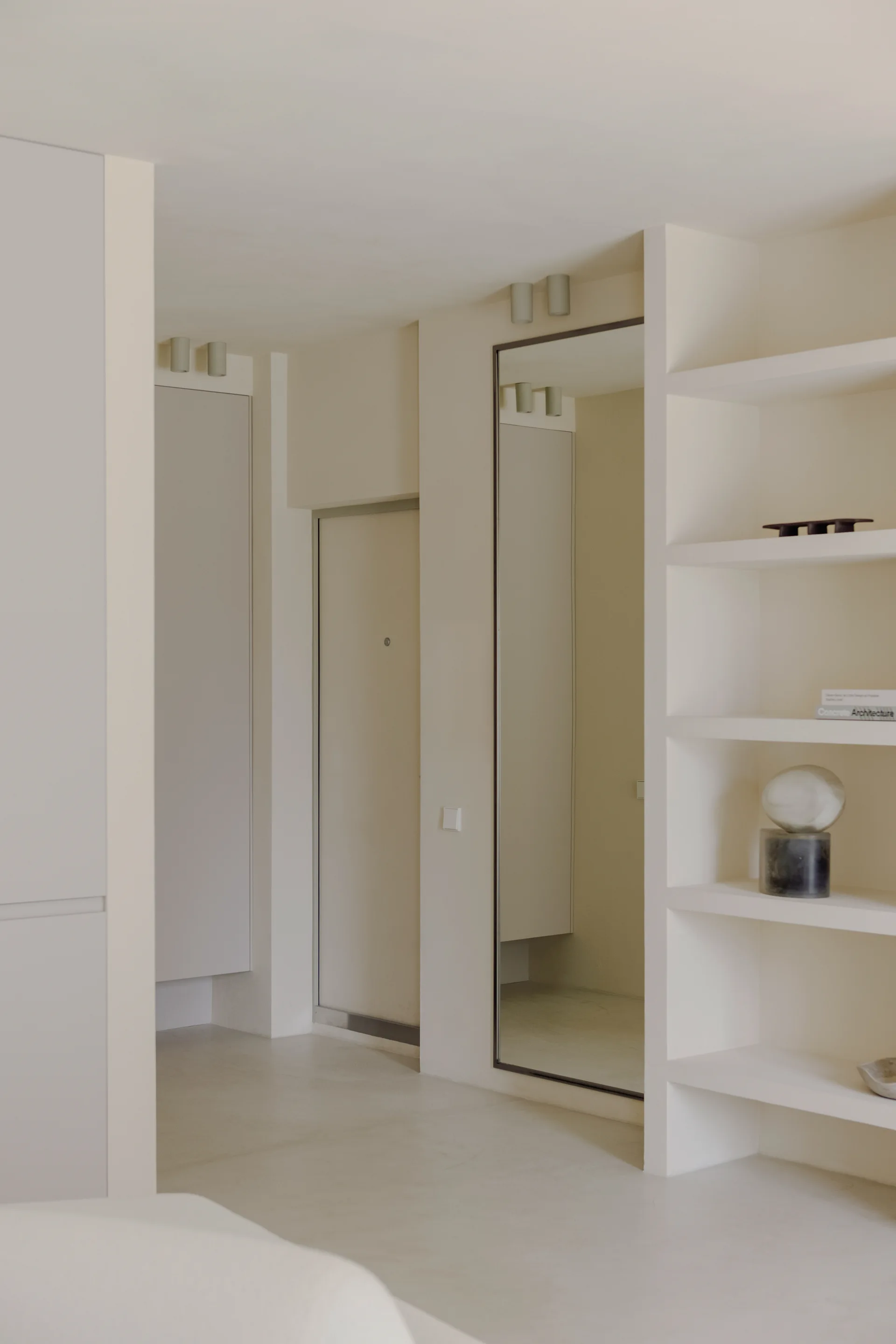A minimalist off-white entryway featuring recessed open shelving, a tall rectangular mirror, and cylindrical ceiling spotlights.