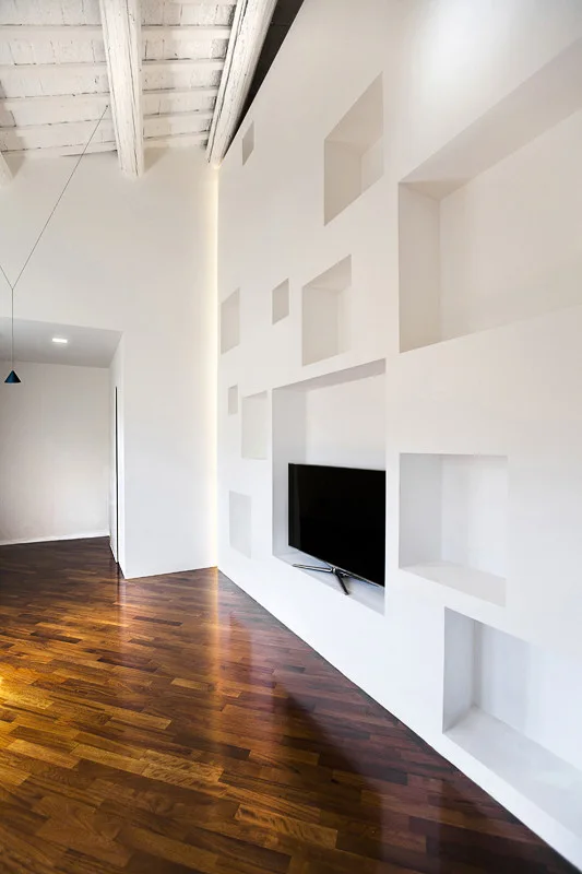 A modern living room features a large white accent wall with recessed shelving niches, a flat-screen television, and dark hardwood floors.