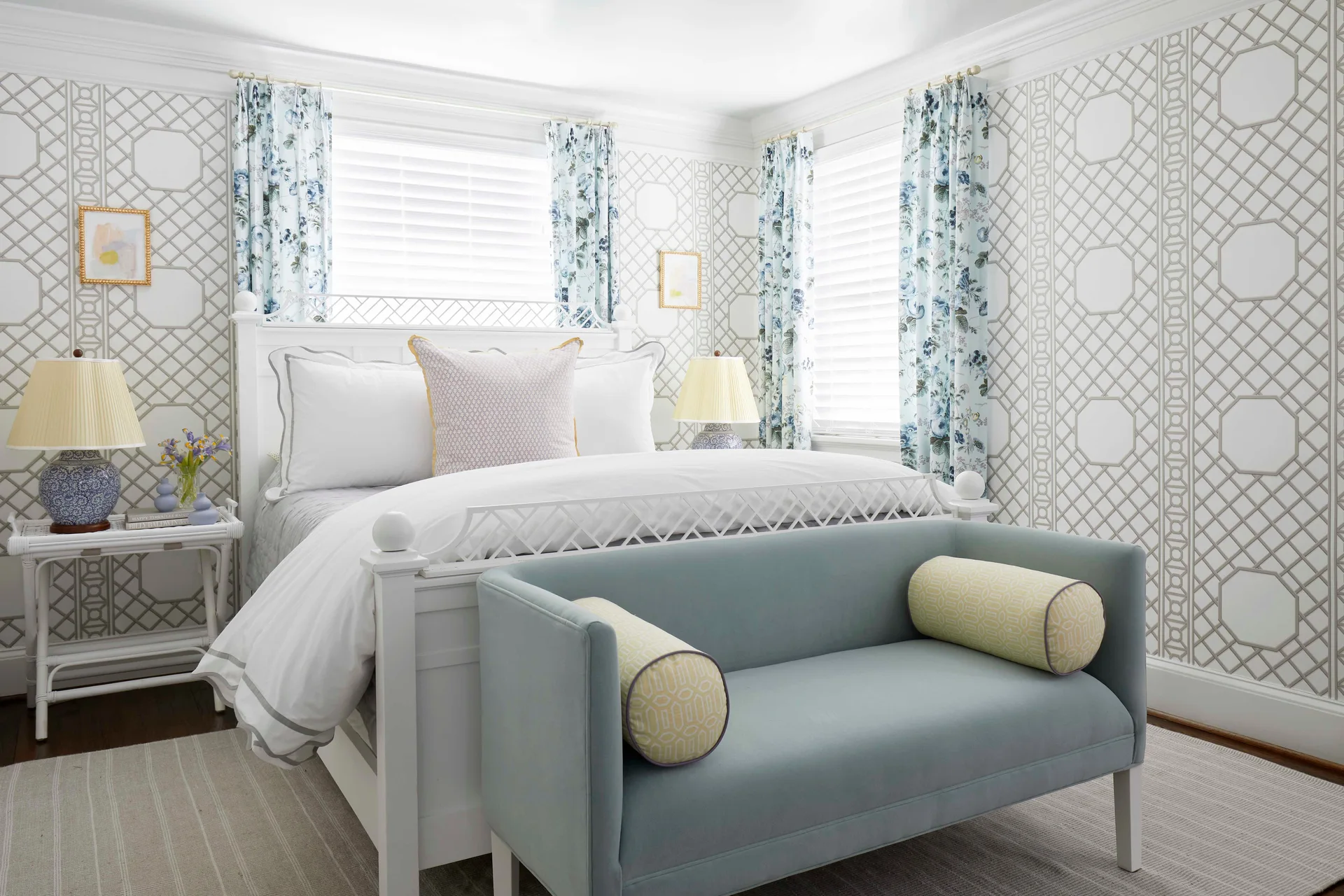 A traditional bedroom features a white bed, a blue upholstered bench, floral drapes, and walls covered in geometric lattice wallpaper.