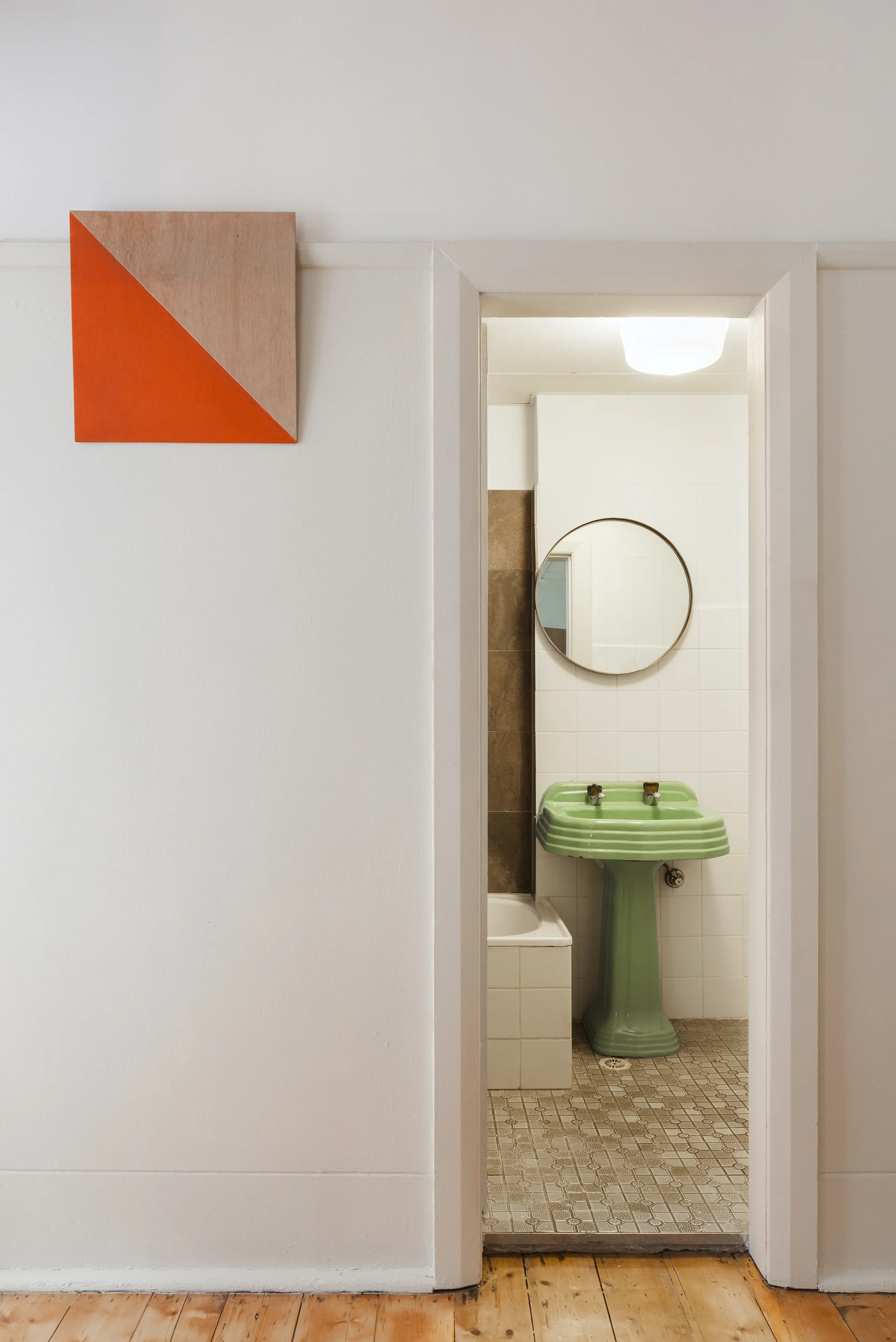 A view through a white doorway showing a vintage bathroom with a green pedestal sink, a round mirror, and patterned floor tile.