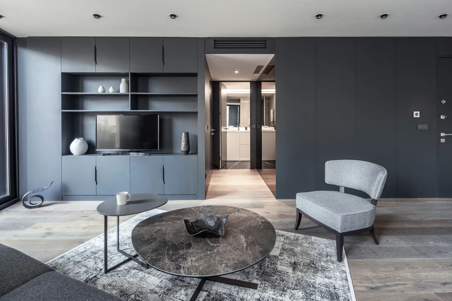 A modern living room features a charcoal grey built-in storage unit with a television, a round stone coffee table, and a grey upholstered armchair.