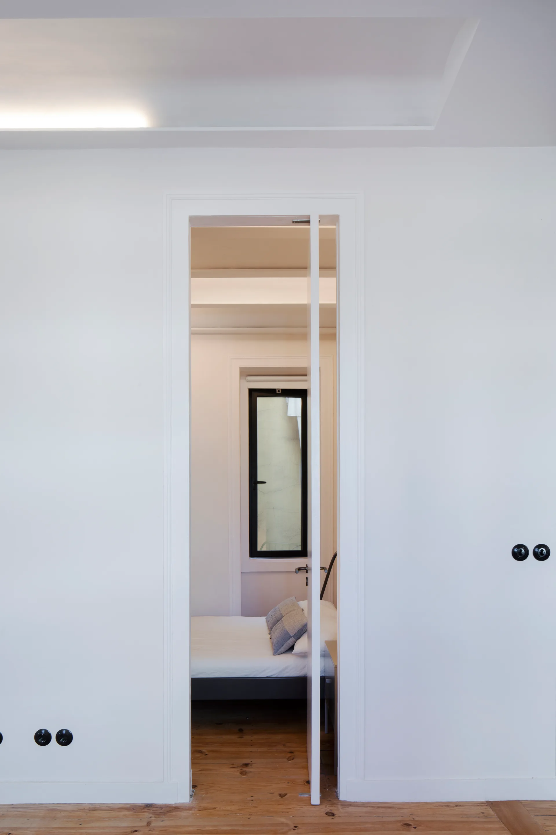 A view through a tall white doorway into a minimalist bedroom with a bed, patterned pillows, natural wood floors, and a narrow black-framed window.