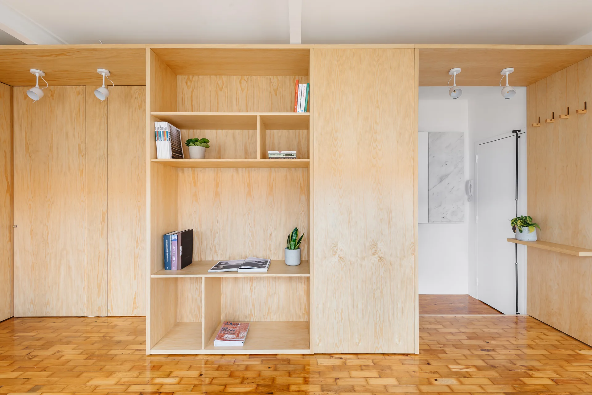 A custom blonde plywood built-in cabinetry unit with open shelves, integrated spotlights, and wooden coat hooks sits on a parquet wood floor.