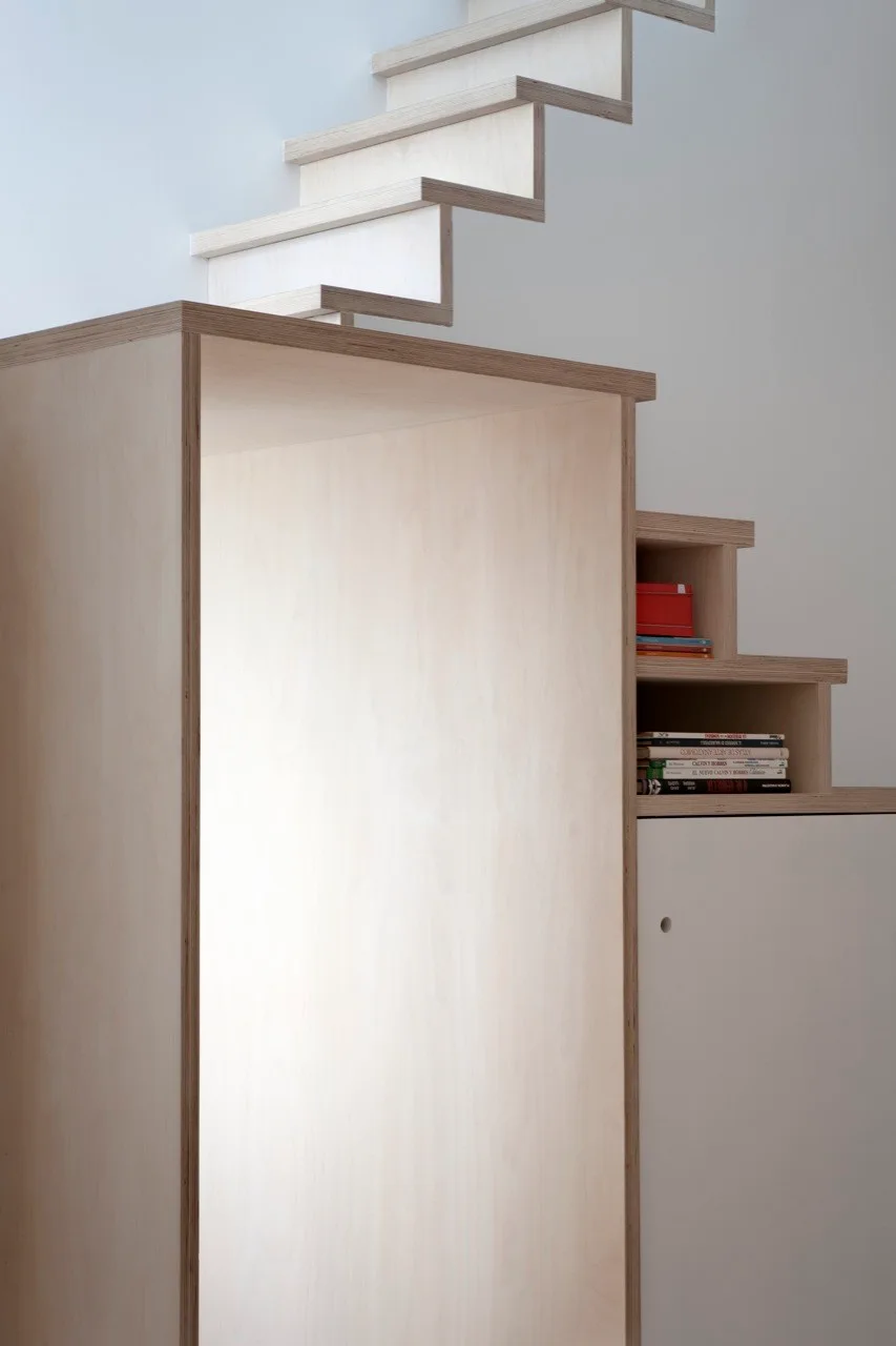 A minimalist light wood plywood staircase features integrated storage cabinets and open shelving displaying books against a white wall.