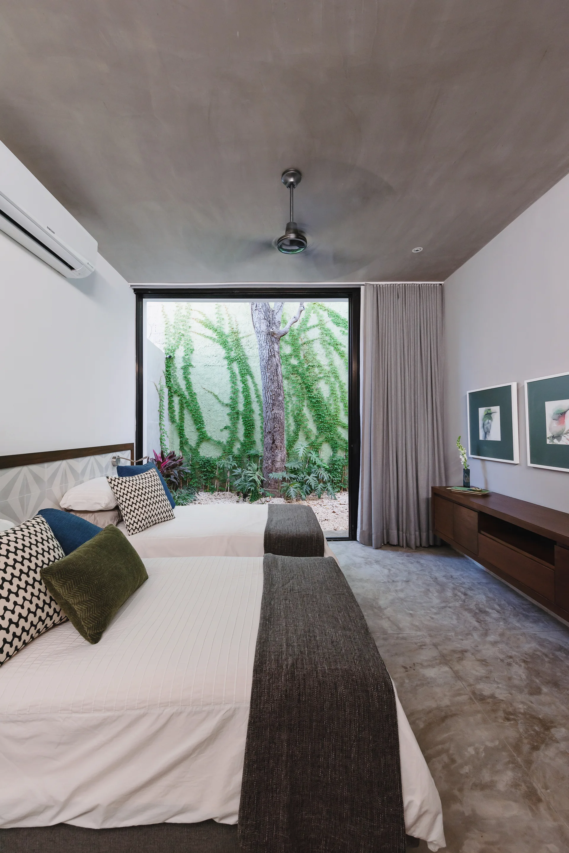 A modern bedroom with two twin beds features a concrete ceiling and floor, with a large sliding glass door opening to a lush garden courtyard.