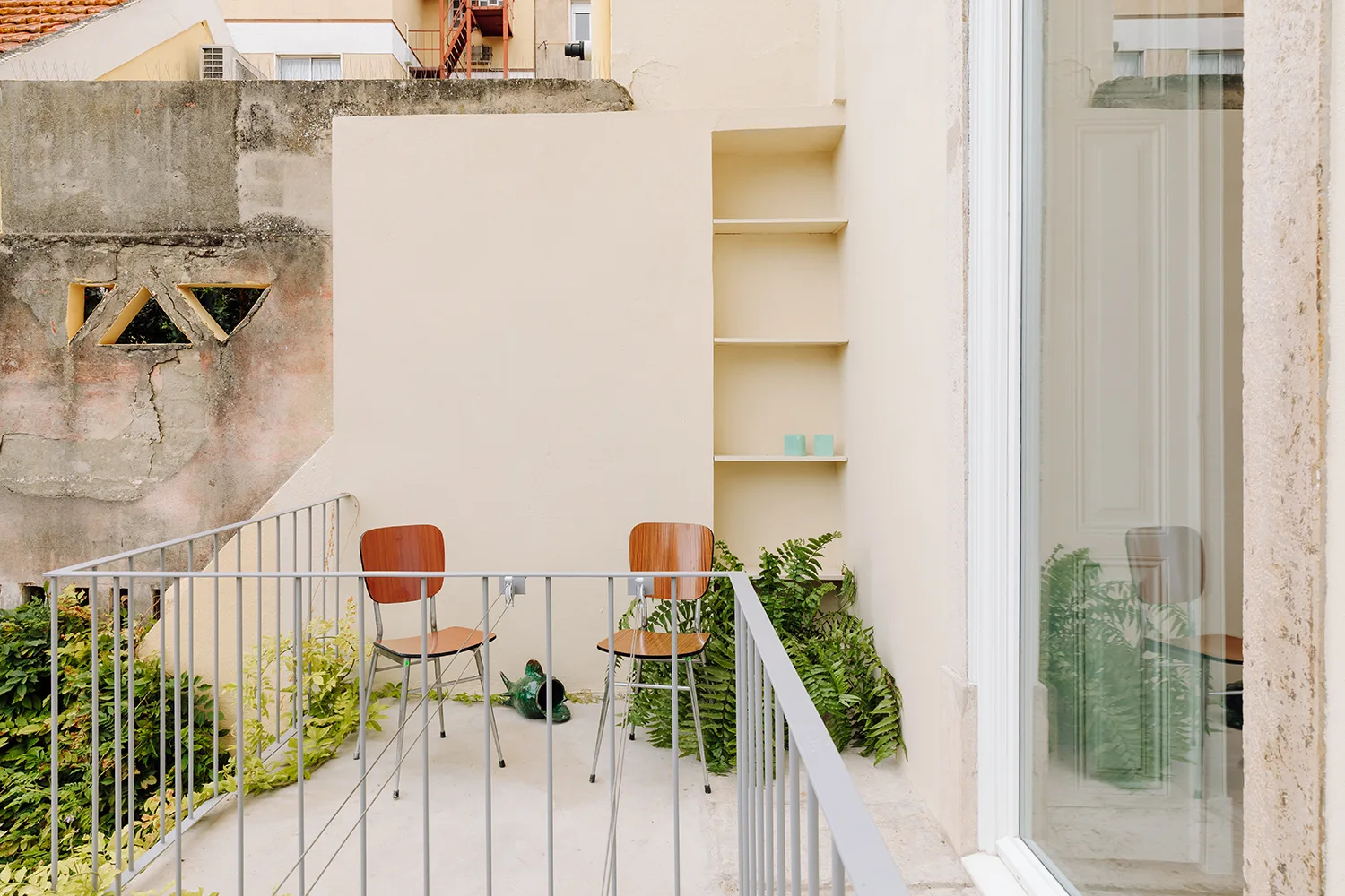 Two mid-century wooden chairs sit on a minimalist beige balcony next to a grey metal railing and a tall recessed shelving unit.