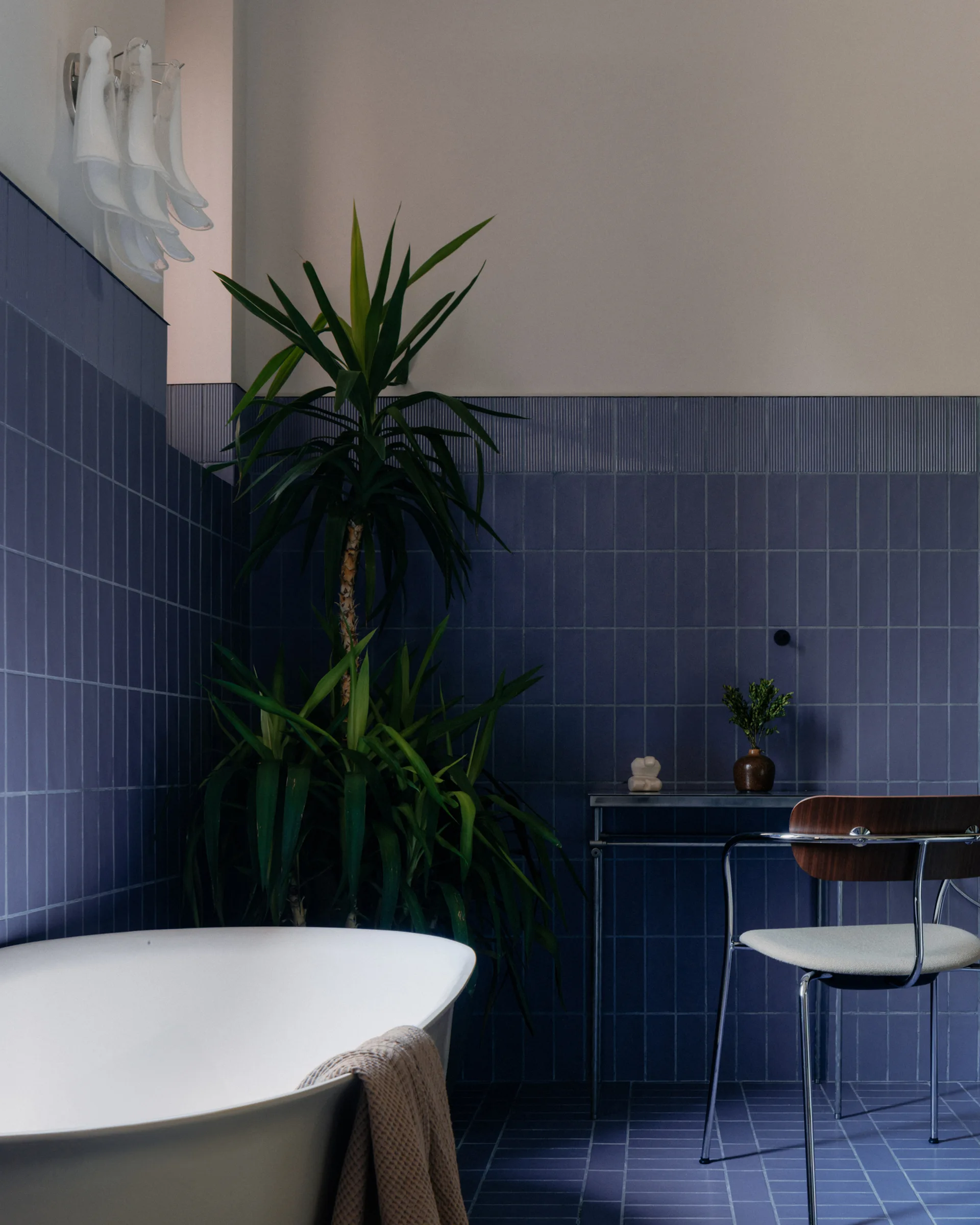 A white freestanding bathtub sits in a corner wrapped in deep blue square tiles, next to a tall potted plant and a metal console table.