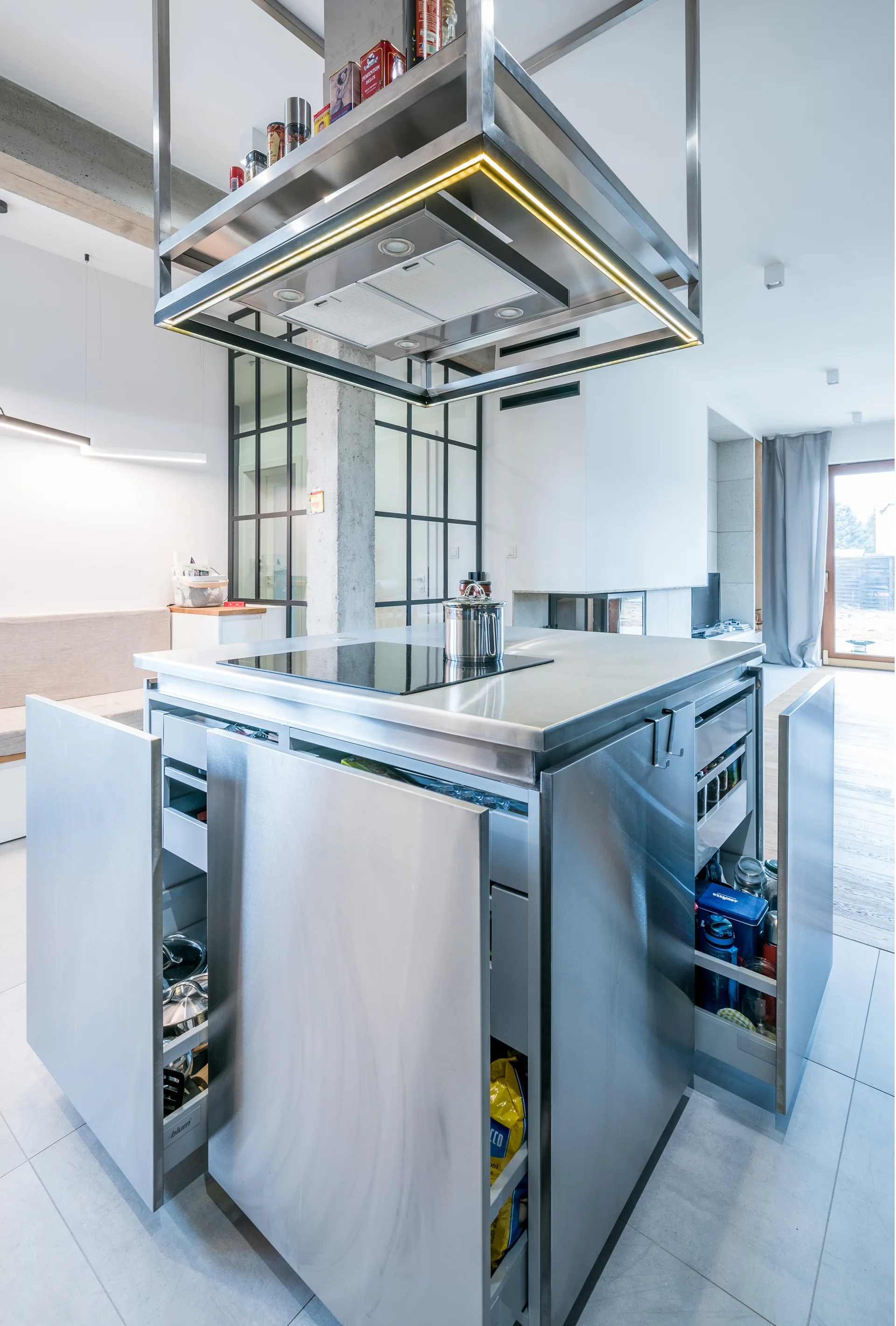 A modern stainless steel kitchen island with open pull-out drawers sits under a suspended metallic range hood with integrated LED lighting.