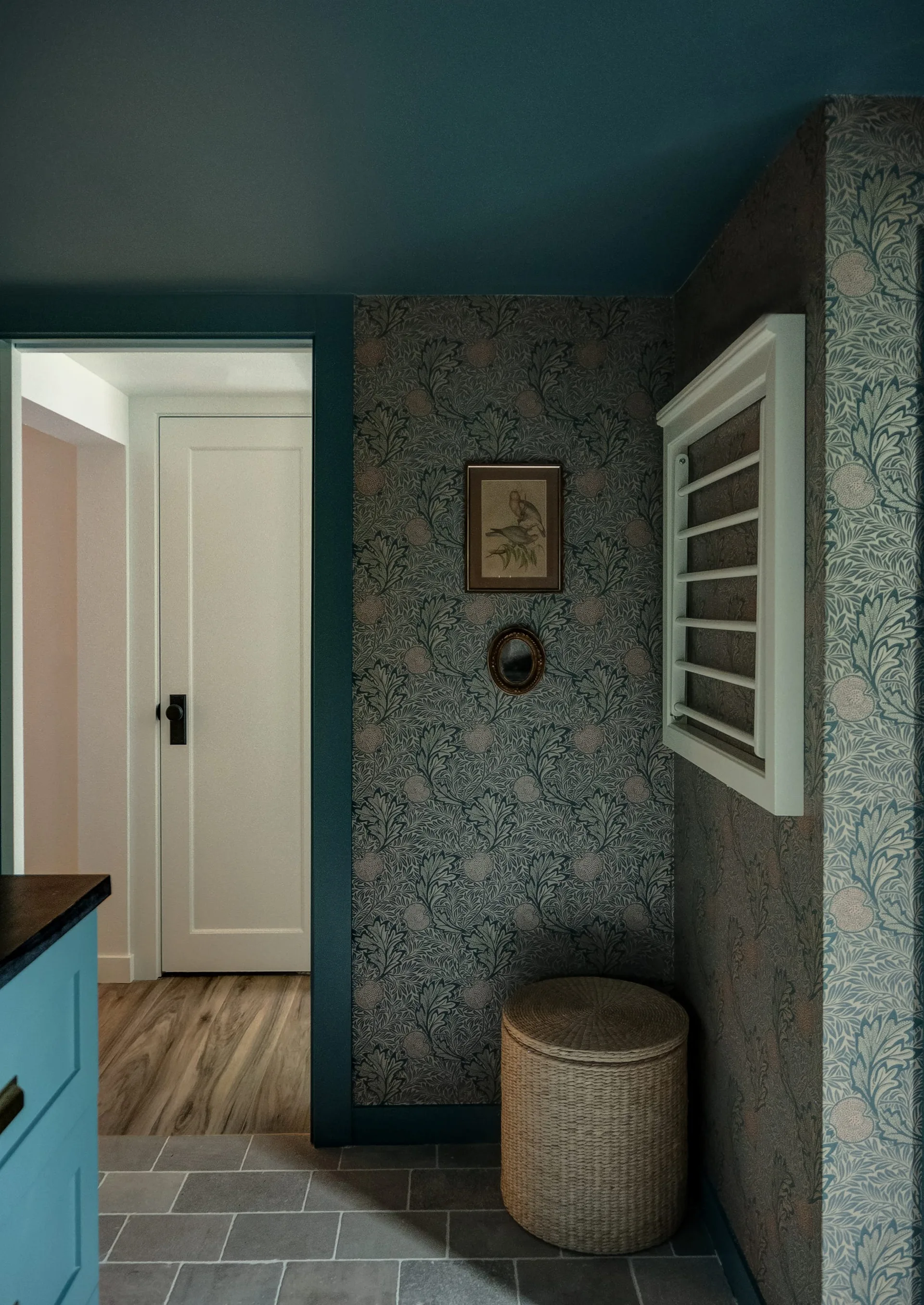 A compact utility room features blue floral wallpaper, a wall-mounted white drying rack, a woven wicker laundry hamper, and a painted blue ceiling.