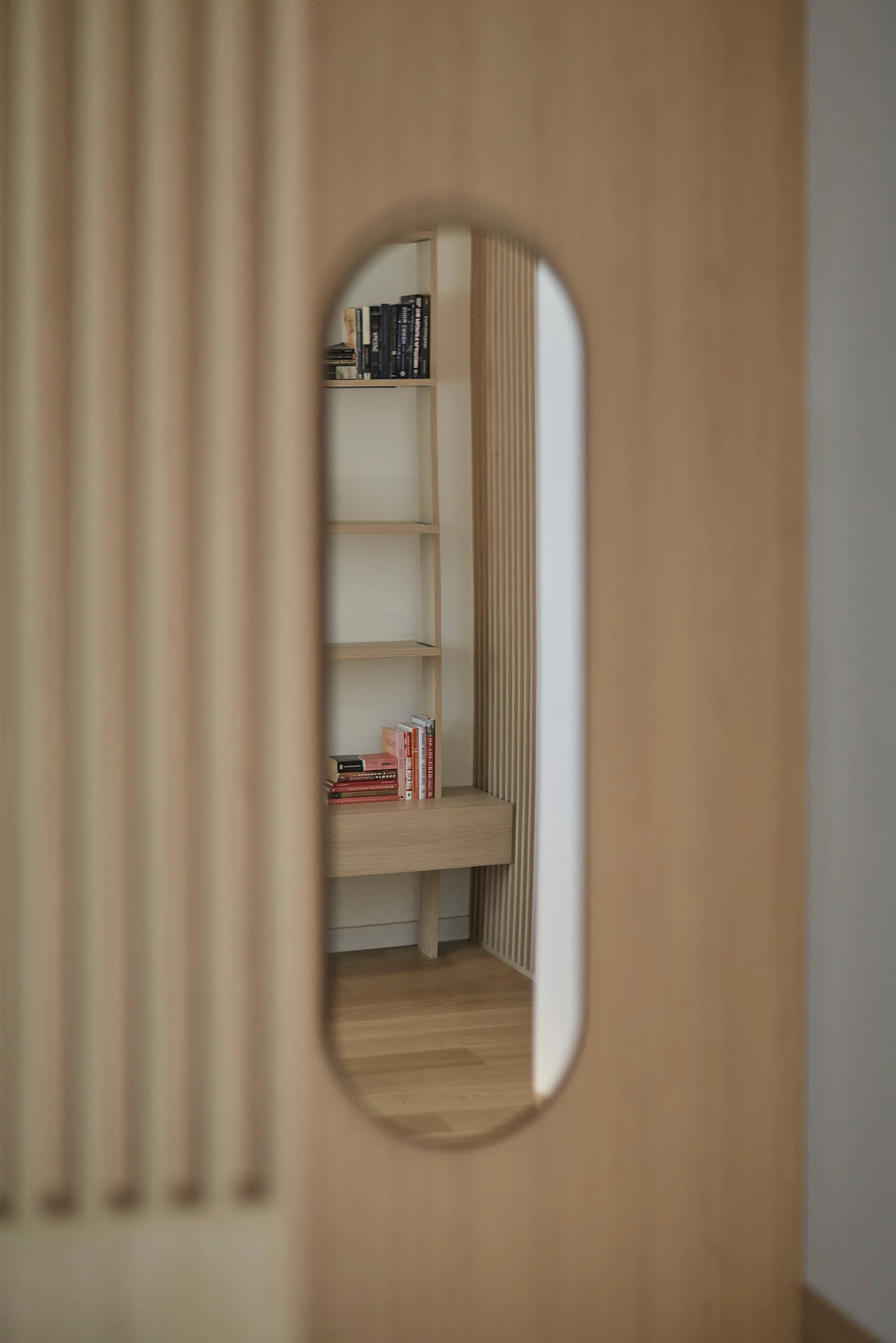 A view through an oval cutout in a fluted light wood partition wall reveals an open bookshelf with books in a minimalist study.