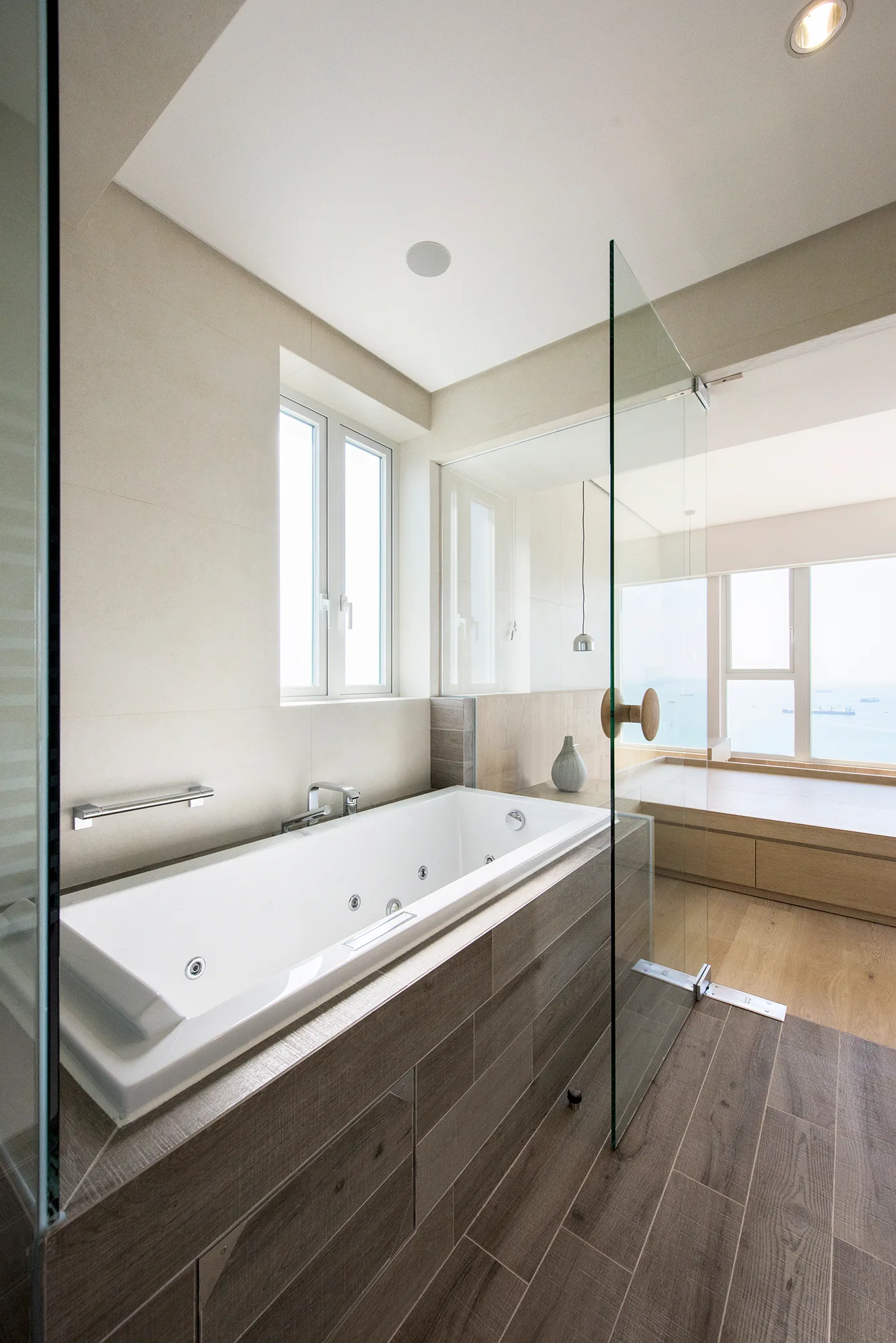 A white jetted bathtub clad in dark wood-look tile is set next to a frameless glass partition in a neutral modern bathroom.