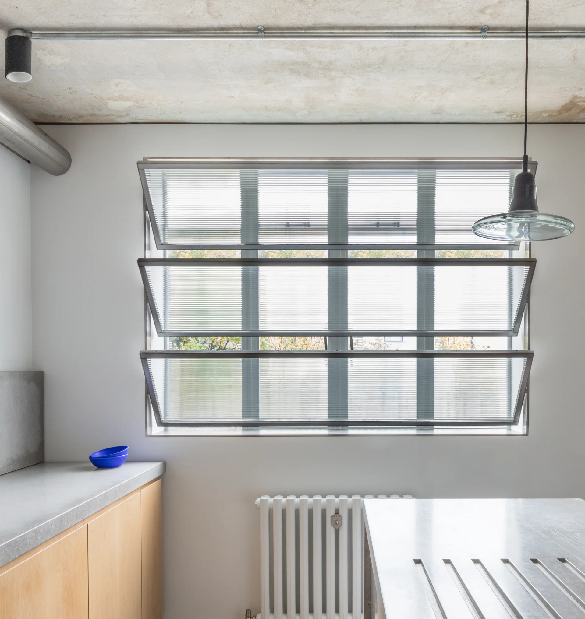 An industrial kitchen features warm wood cabinetry, grey concrete countertops, an exposed concrete ceiling, and a ribbed glass awning window.