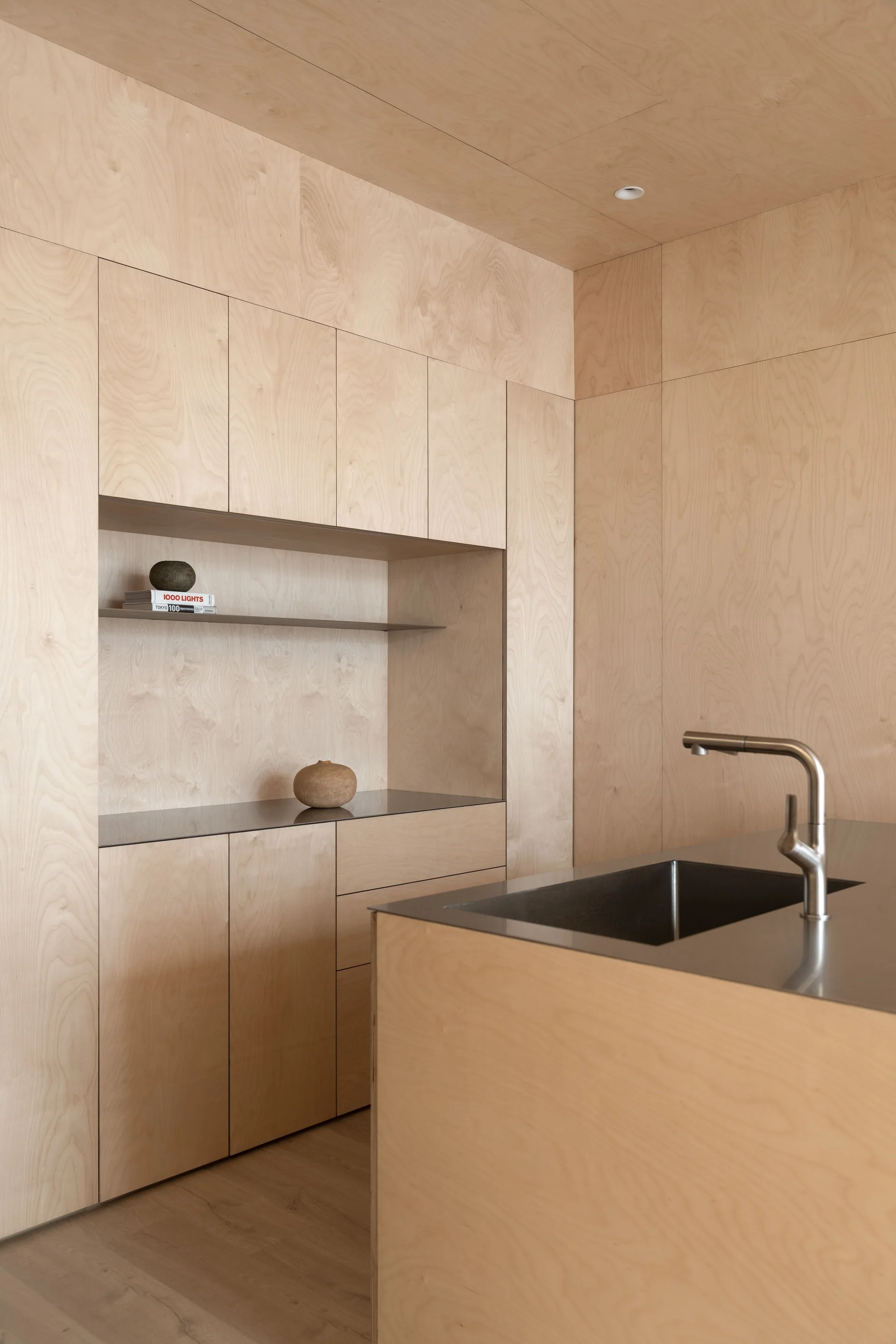 A minimalist kitchen features custom light-colored plywood walls, built-in cabinetry with a recessed shelving niche, and a stainless steel island sink.