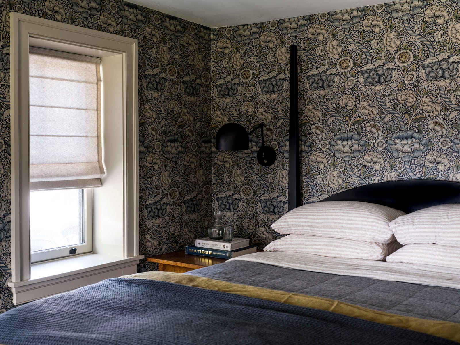 A bedroom corner features botanical wallpaper, a bed with gray bedding, a black wall sconce, and a recessed window with a beige Roman shade.