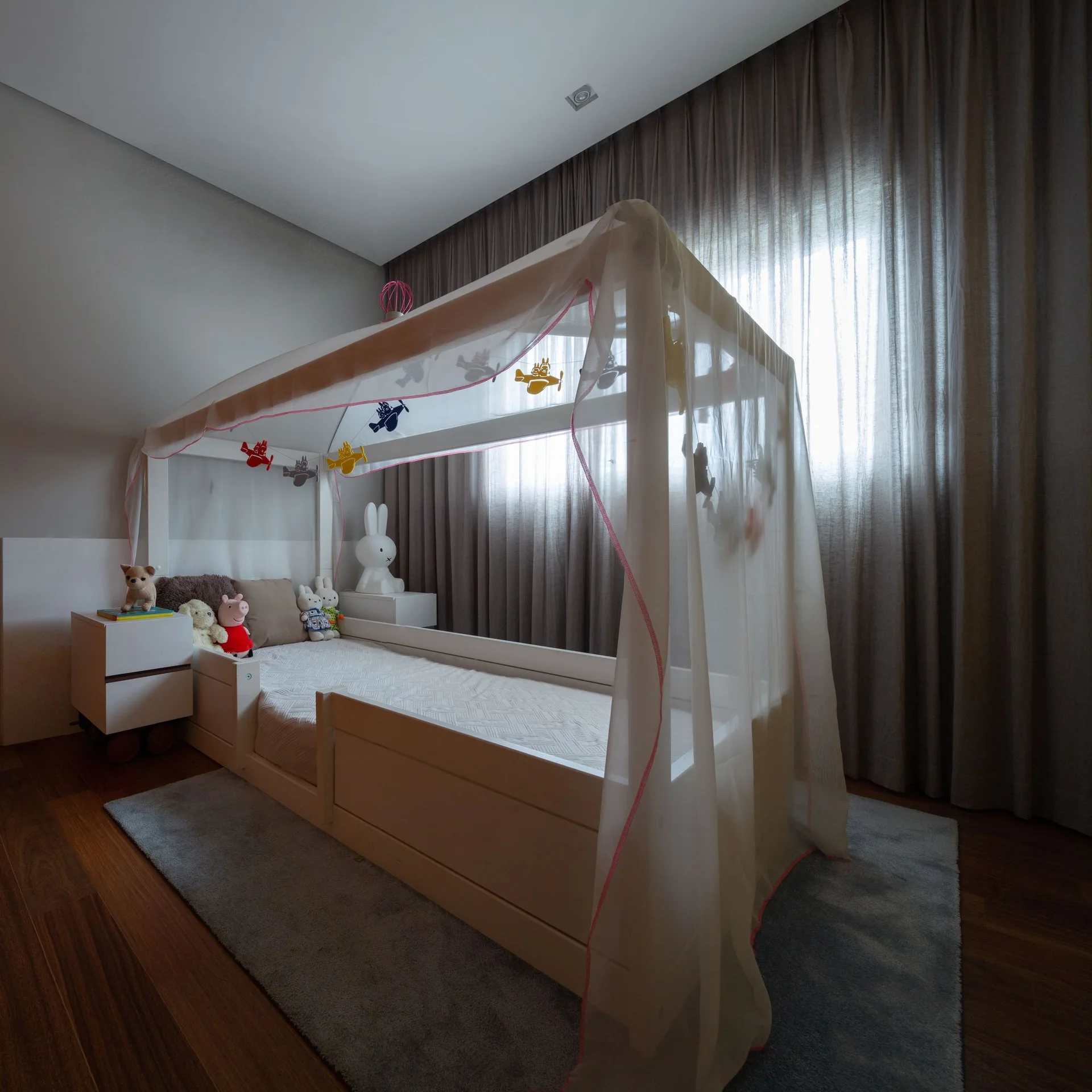 A childrens bedroom features a white wooden canopy bed with sheer draping, a gray area rug, and floor-to-ceiling gray curtains.