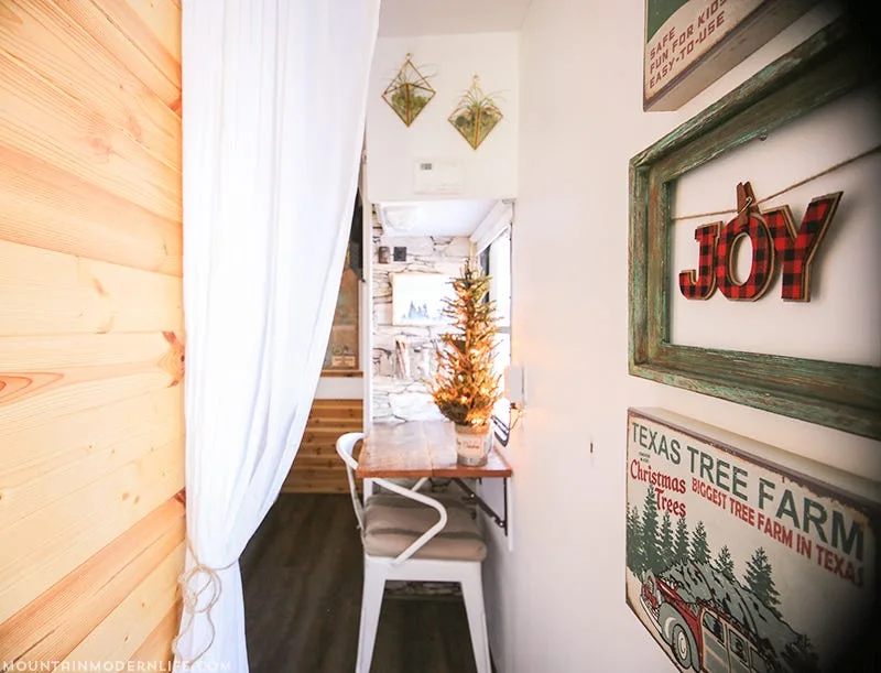 A narrow camper interior features a wall-mounted wooden table, a white metal chair, and rustic holiday wall decor beside a white curtain.