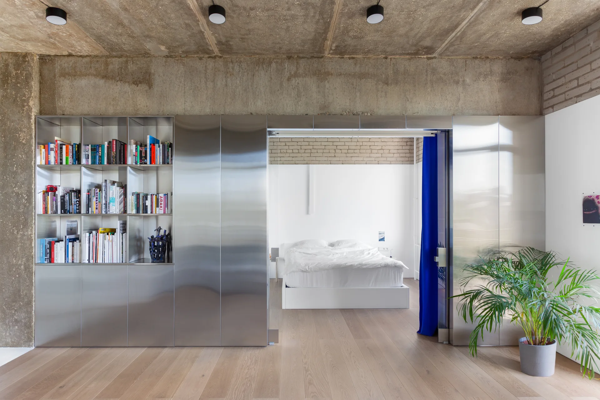A modern bedroom visible through a brushed metal partition wall with bookshelves next to an exposed concrete ceiling and a bright blue curtain.
