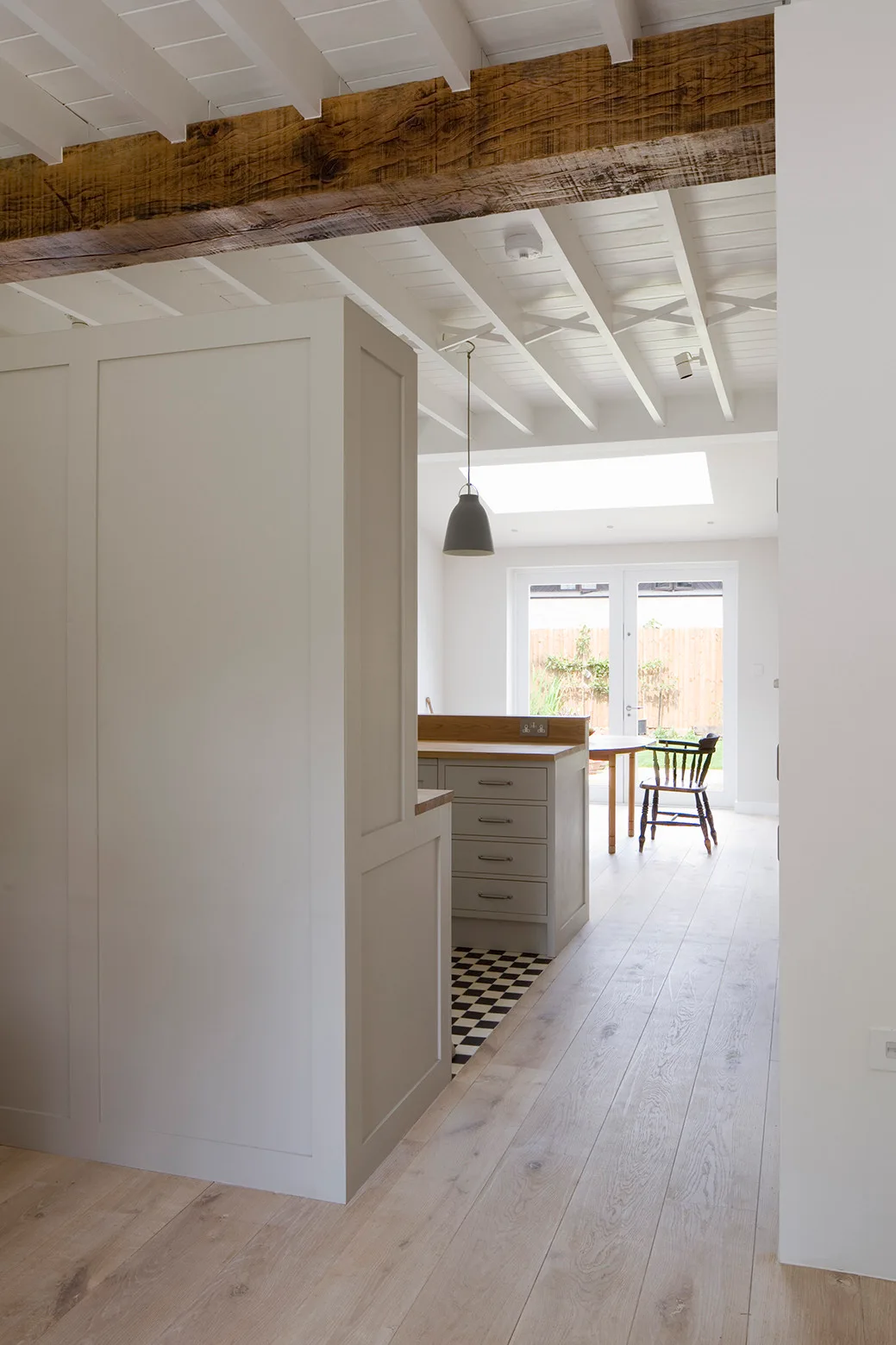 An off-white kitchen cabinet divider and light wood floors that transition to checkerboard tiles beneath an exposed ceiling beam.