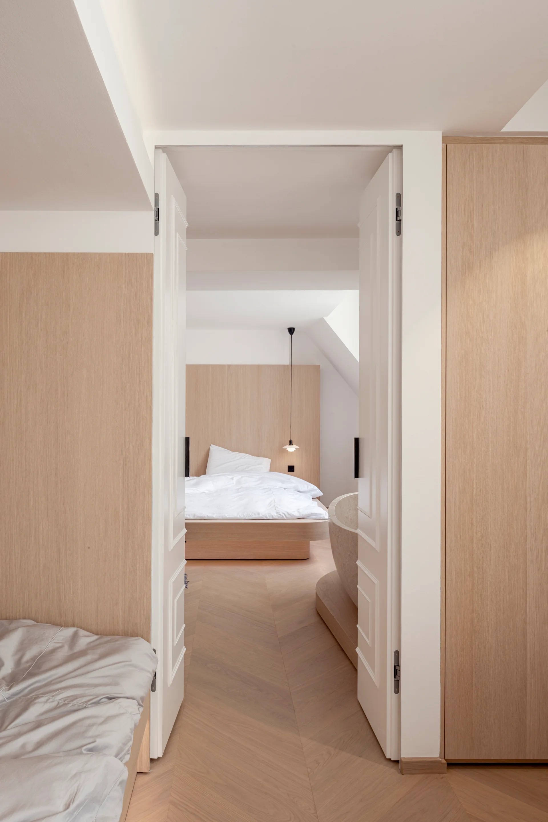 A view through open white double doors into a minimalist bedroom featuring a bed, light wood paneling, and a stone freestanding bathtub.