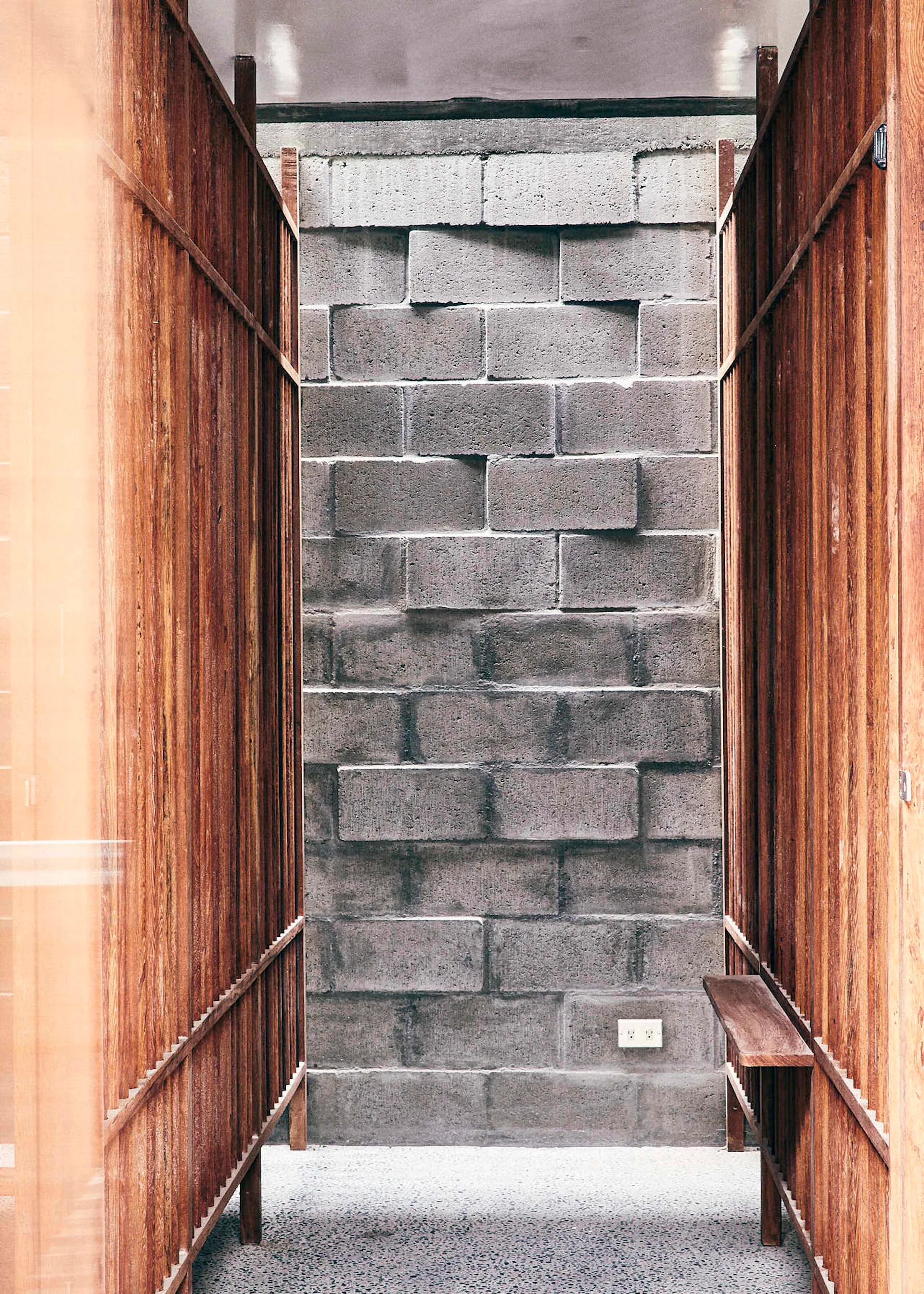 A narrow corridor features tall vertical wooden slat panels flanking a central path that leads to an exposed grey concrete block wall.