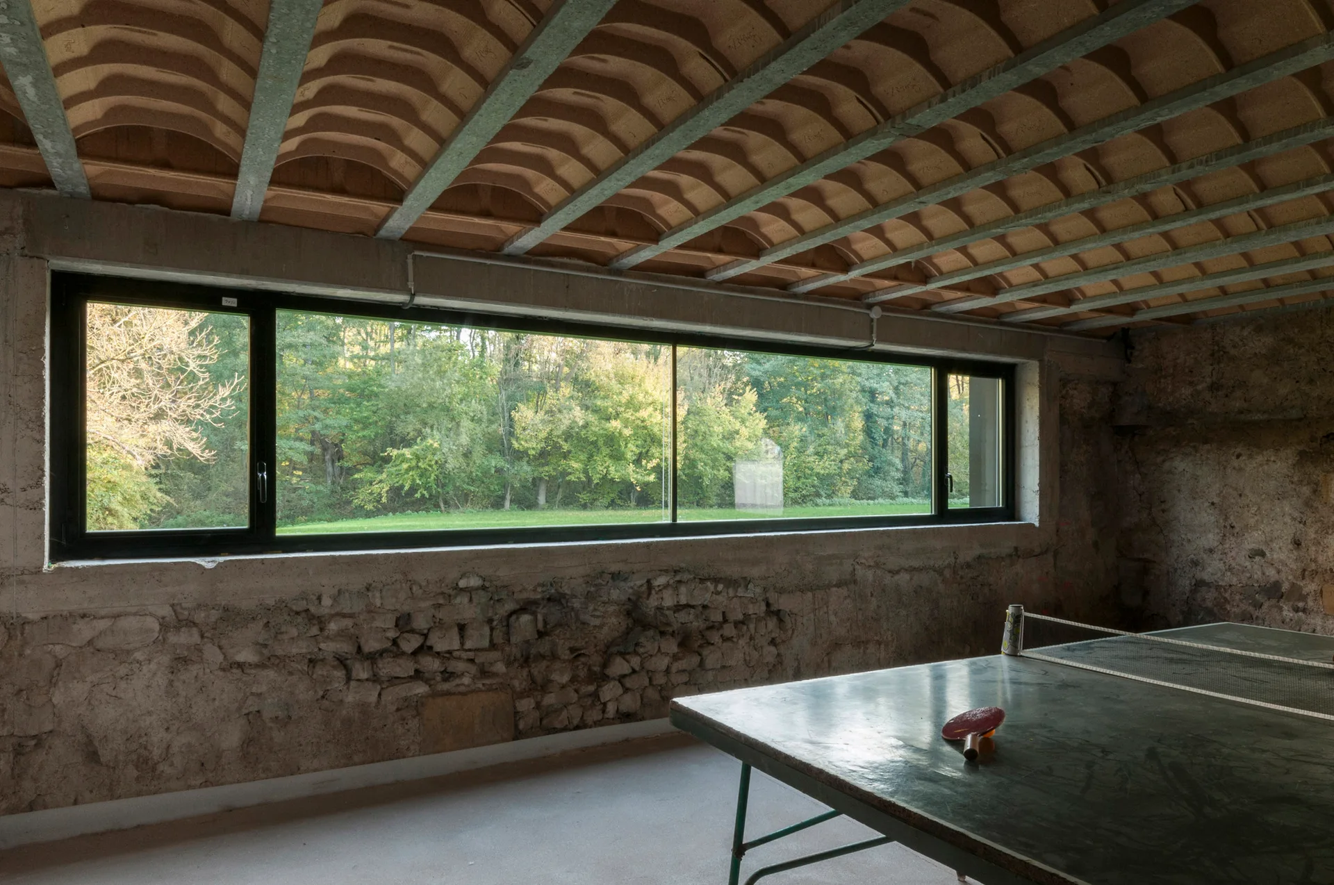 A rustic recreation room with a ping pong table, an exposed stone wall, and a large horizontal window showcasing a forest view.