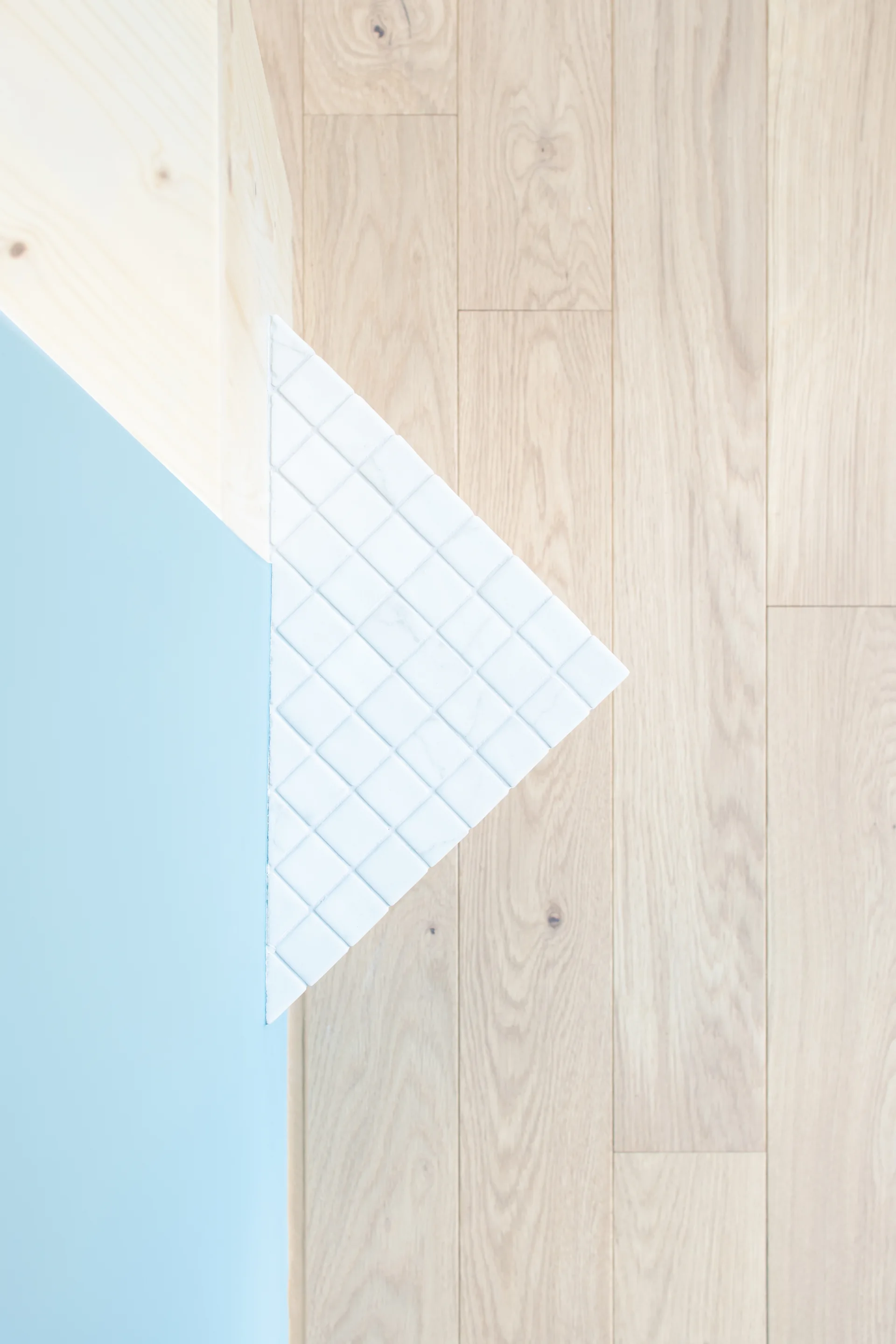 A close-up of a geometric structural corner featuring a blue painted wall, a light wood surface, and white square mosaic tiles over wood plank flooring.