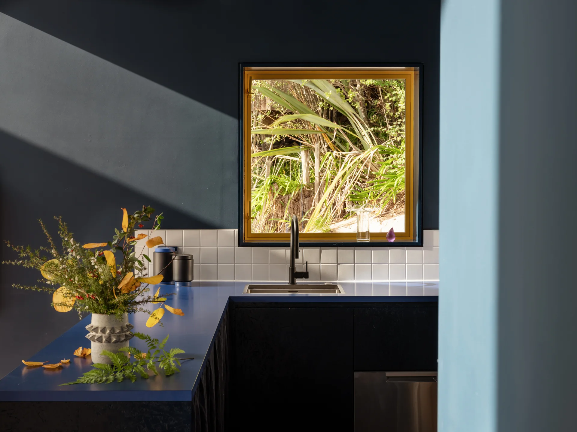A modern kitchen corner featuring a blue countertop, a dark faucet, white square tiles, and a square window with a yellow frame.