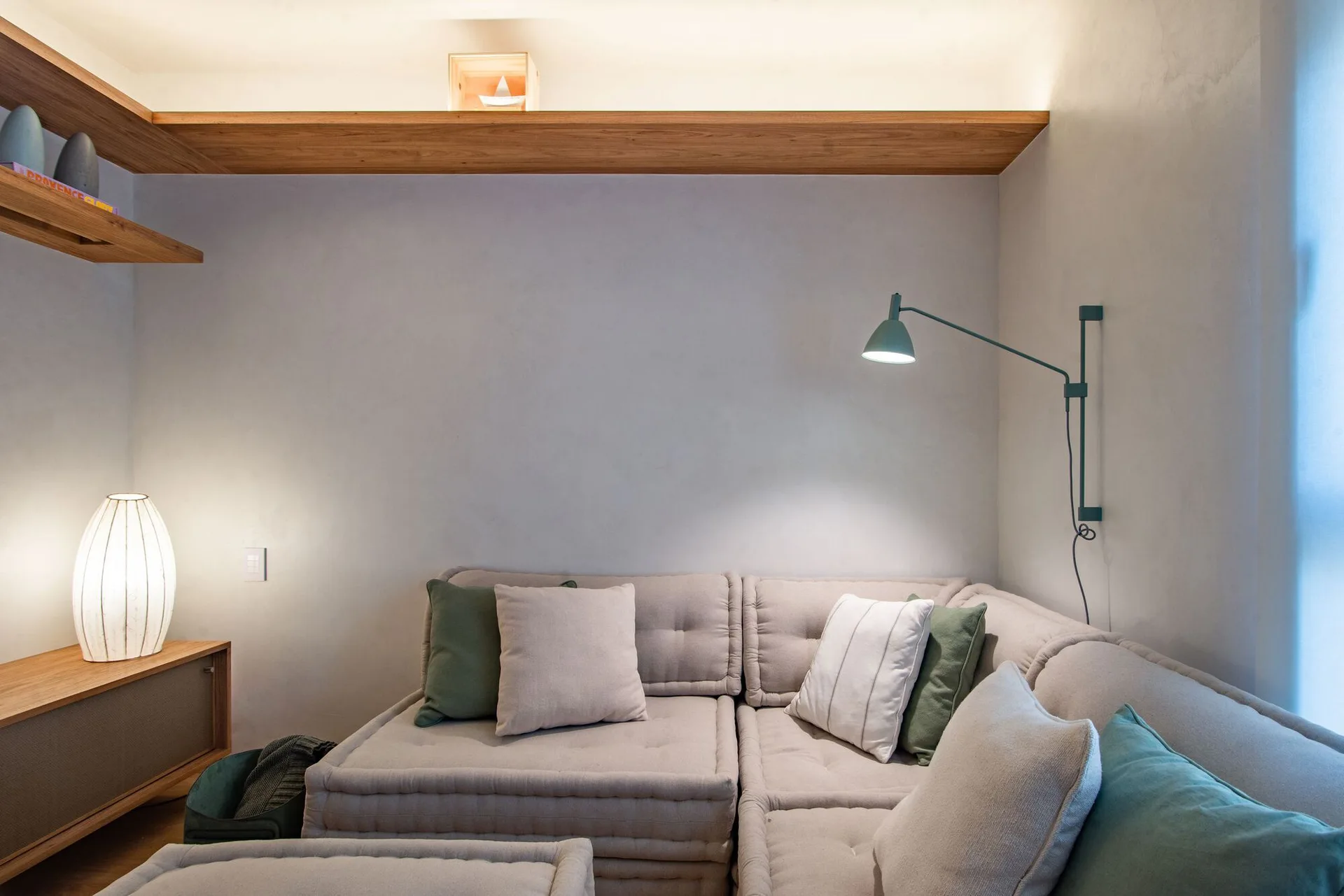 A low modular beige sofa sits in a living room corner under a wooden floating shelf with integrated lighting and a green swing-arm wall sconce.