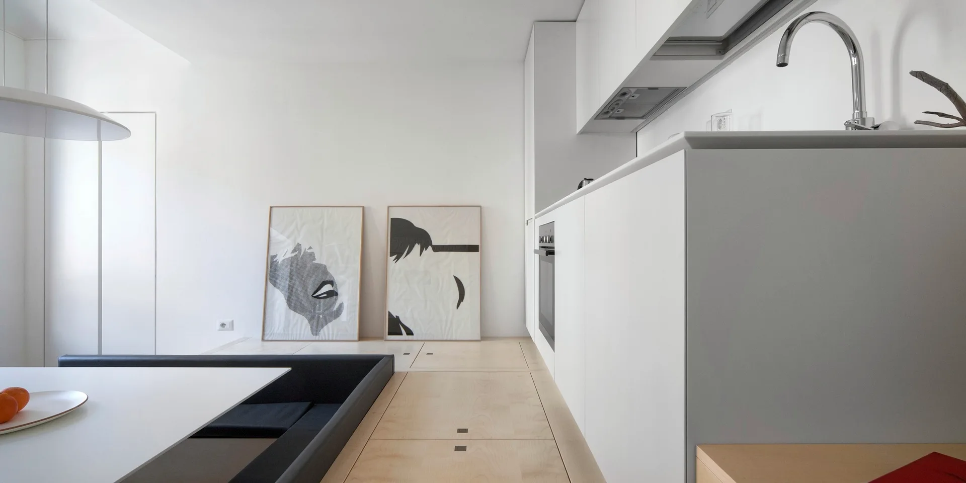 A minimalist white kitchen featuring seamless flat-front cabinets and a raised light wood platform floor with a sunken dining area.