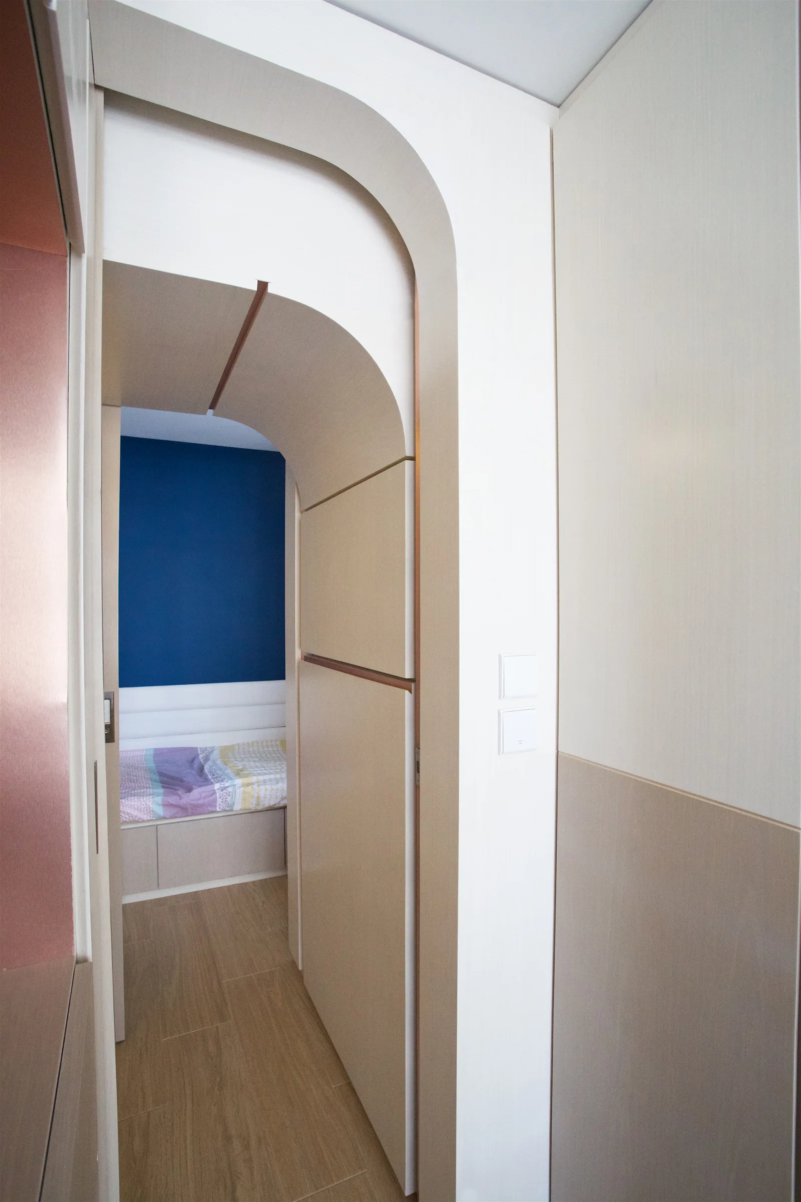 A light beige arched doorway built into cabinetry leads to a bedroom featuring a deep blue accent wall and a built-in bed platform.