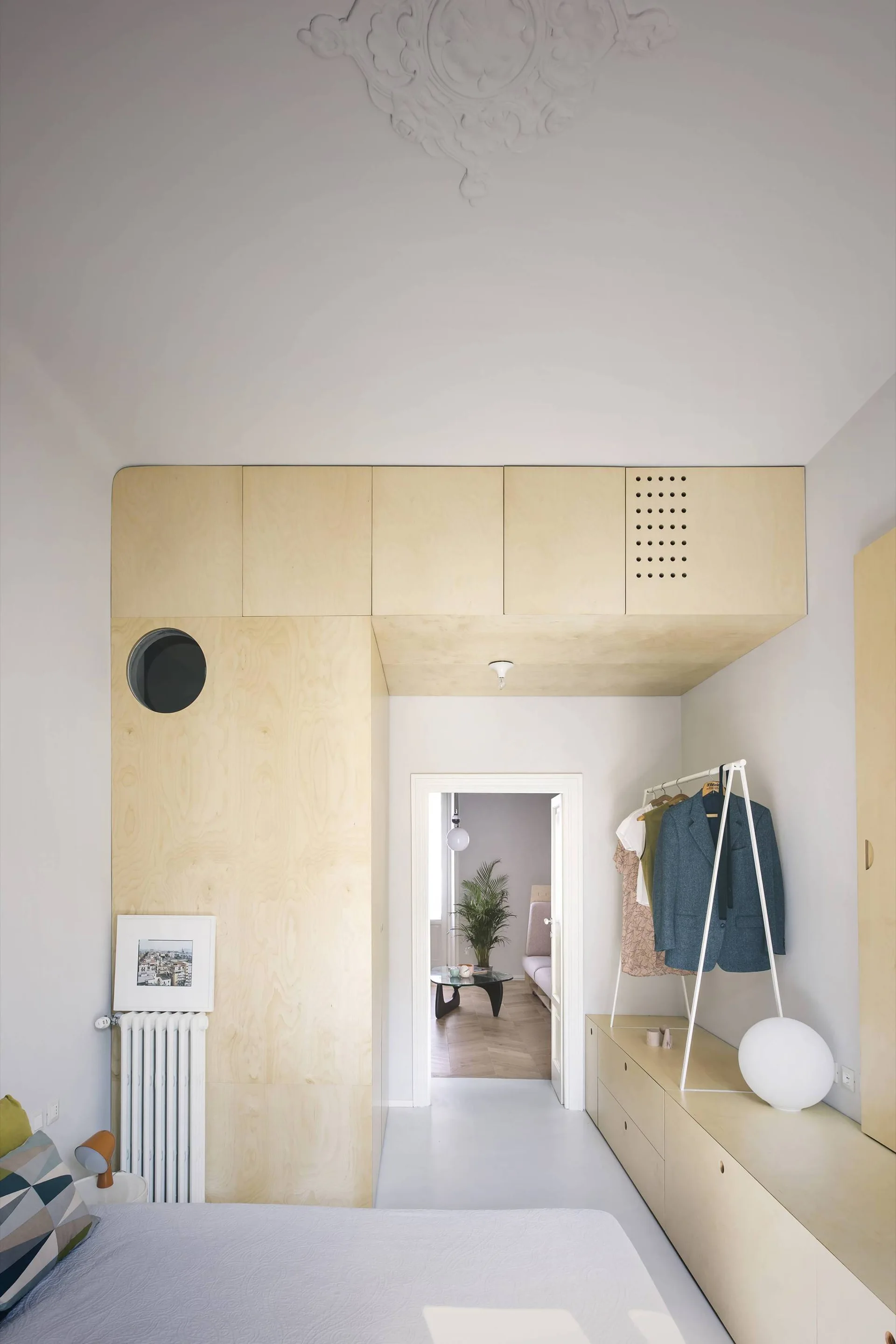 A minimalist bedroom features light plywood cabinets built around a doorway, a white clothing rack, and an ornate ceiling medallion.