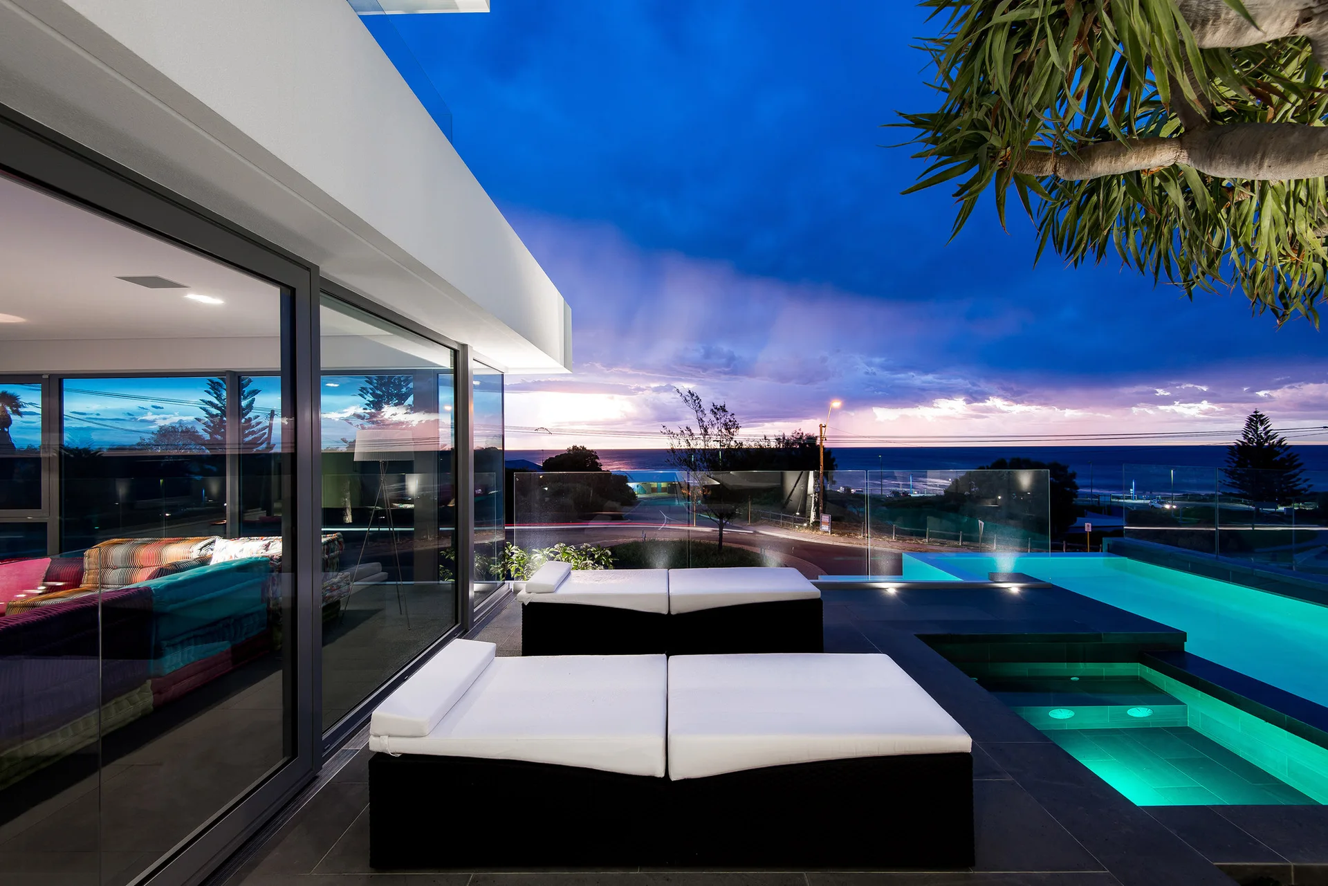 Two white chaise lounges sit on a dark-tiled patio beside an illuminated swimming pool with glass railings overlooking the ocean.