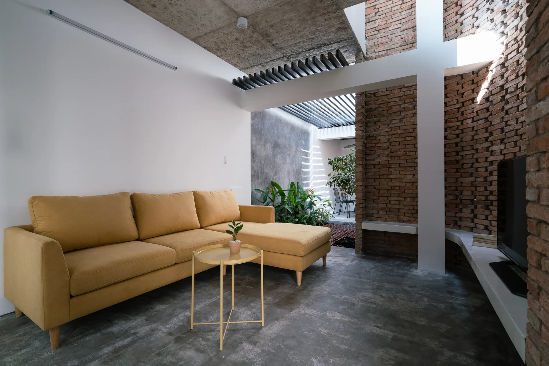 A mustard yellow sectional sofa sits in a modern living room with concrete floors, a raw concrete ceiling, and a curved exposed brick wall.