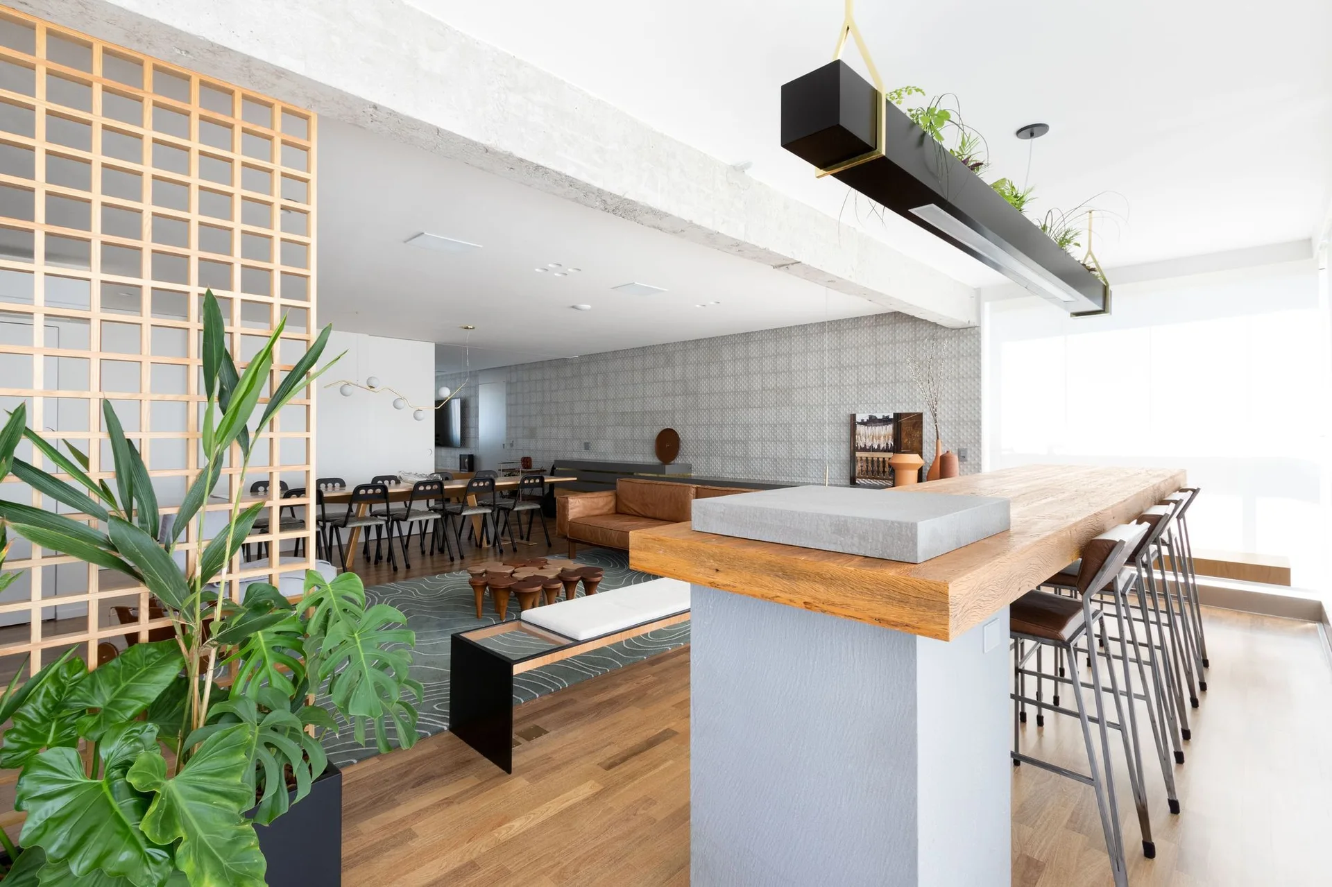 A modern open-plan living area featuring a wooden kitchen island with bar stools, a leather sofa, a wood grid divider, and an exposed concrete ceiling beam.