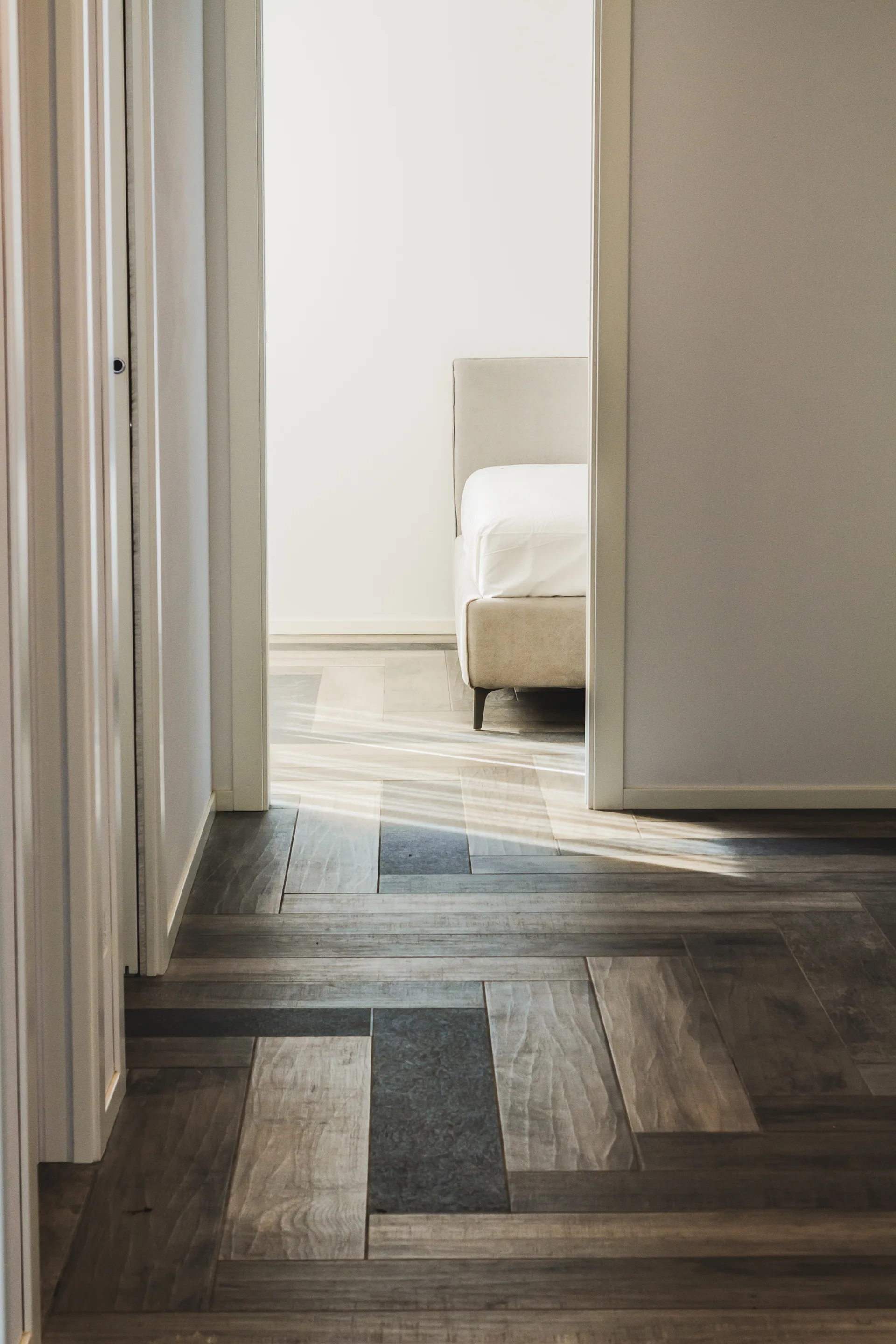 A view through an open doorway shows a neutral-toned bedroom with a beige bed and light streaming onto multi-toned wood herringbone floors.