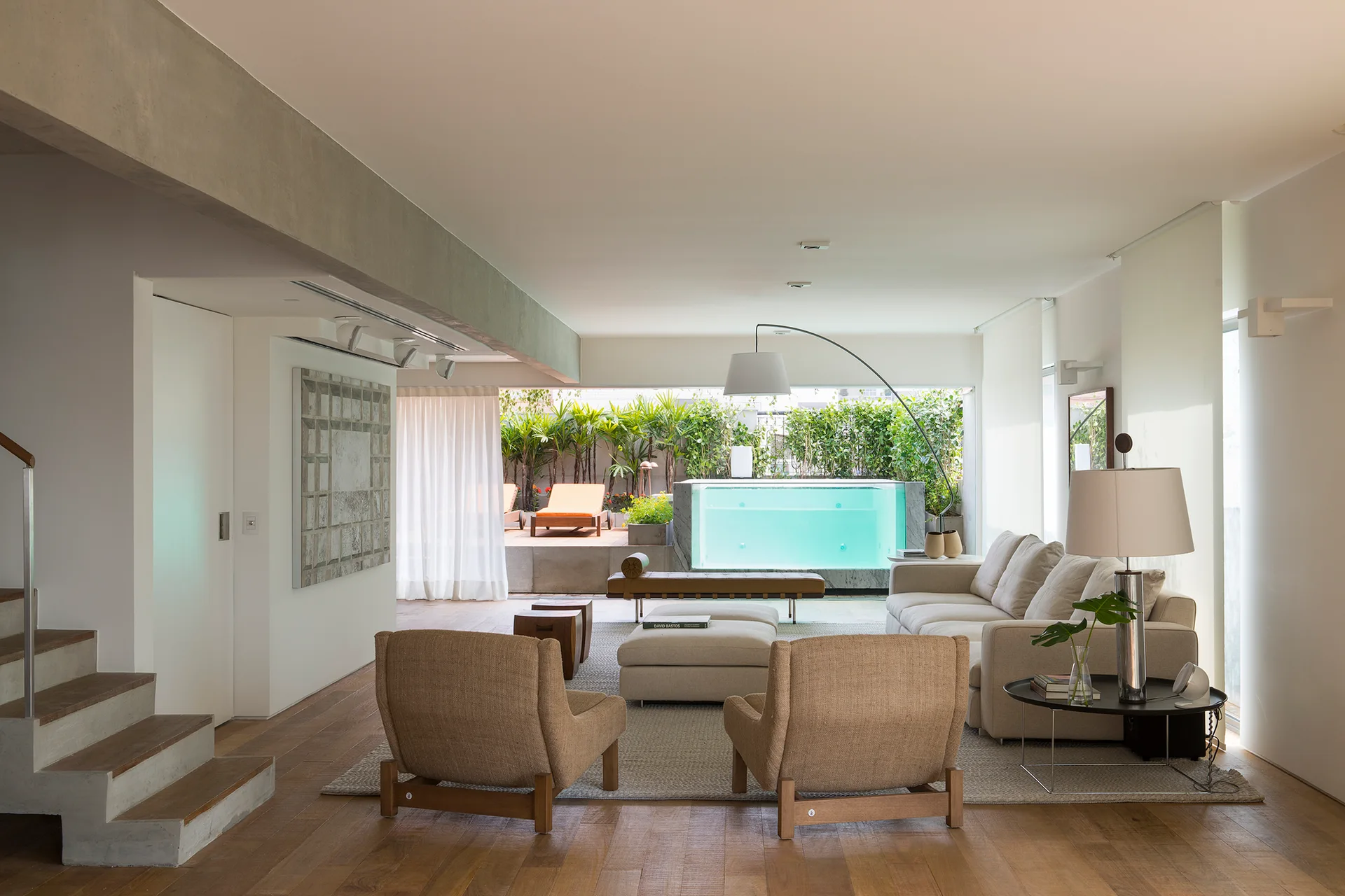 A minimalist living room with a beige sectional sofa, woven armchairs, an exposed concrete beam, and a view of an illuminated terrace pool.