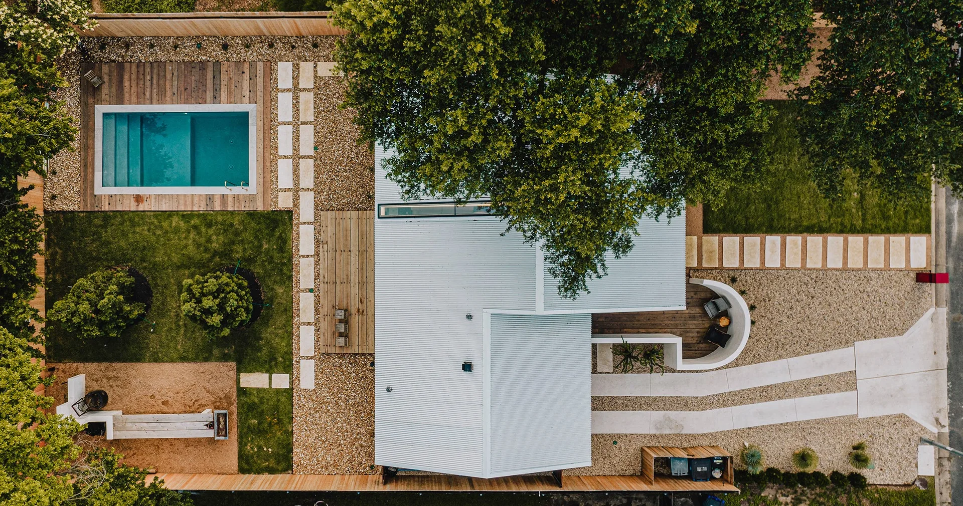 An aerial drone view of a modern backyard featuring a corrugated metal roof house, rectangular swimming pool, wood decks, and stone paver paths.