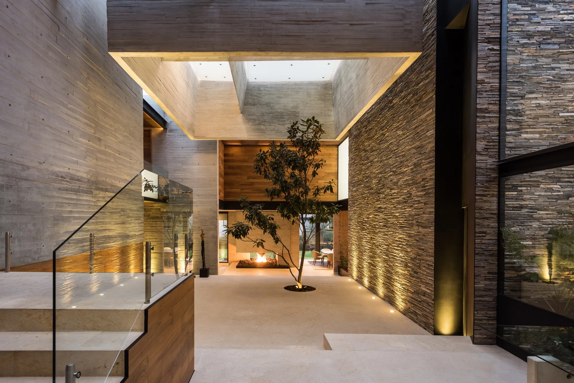 A modern architectural interior features a central live tree under a skylight, surrounded by board-formed concrete and stacked stone walls.
