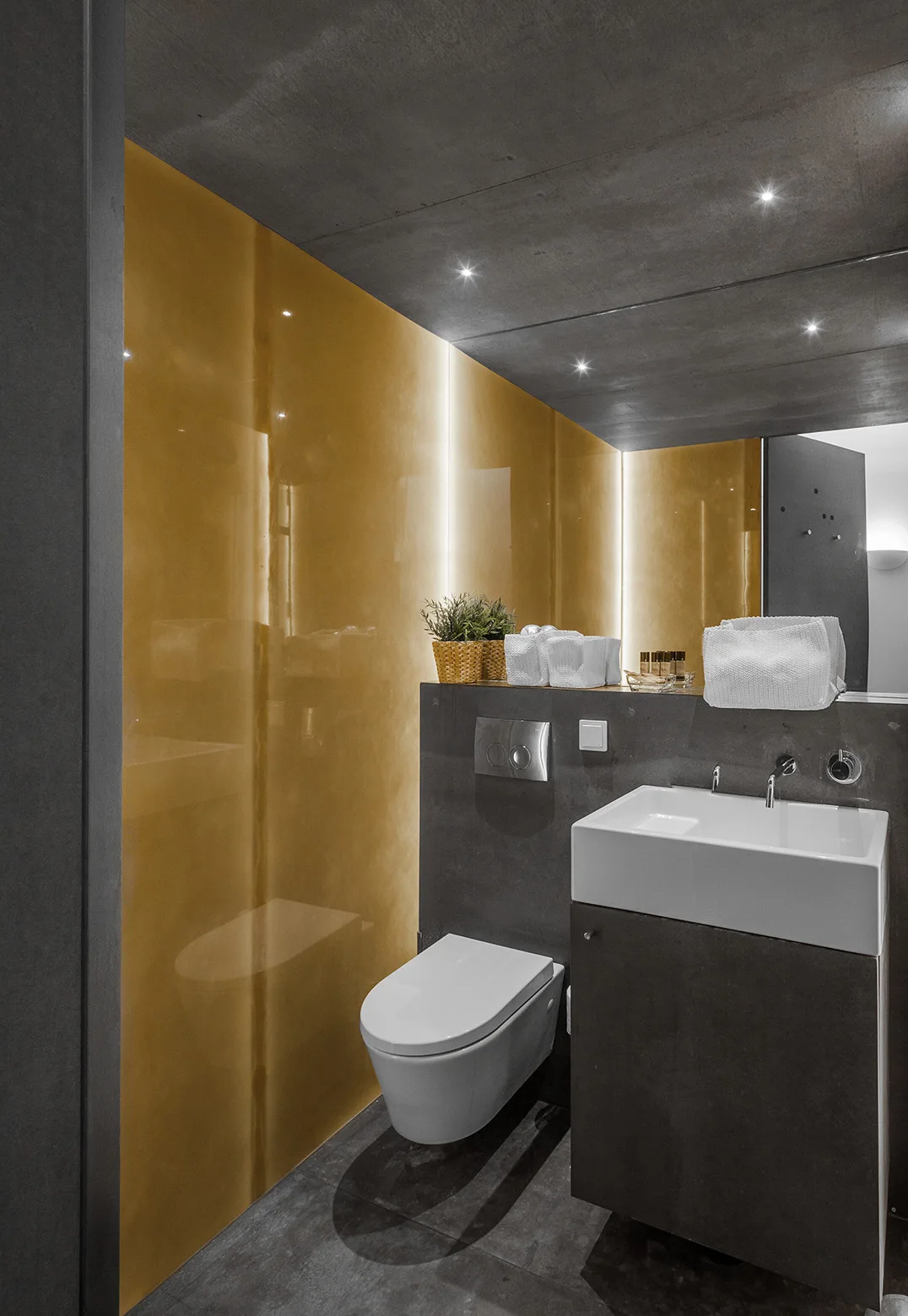 A modern bathroom features a reflective gold wall panel, a wall-mounted toilet, a white vessel sink on a dark grey vanity, and dark concrete ceilings.