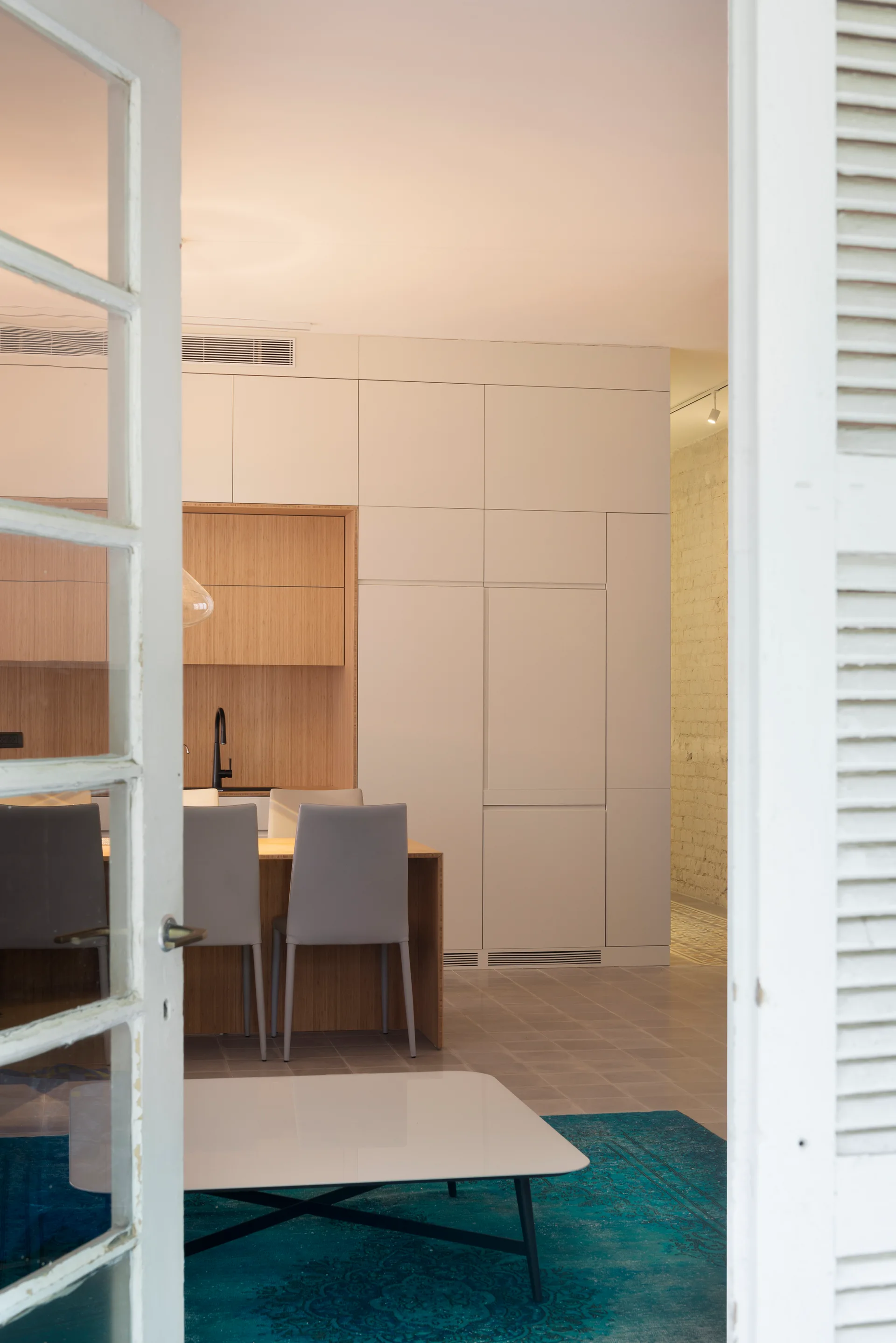 A view through open white shuttered doors into a modern kitchen with flat-panel white cabinetry, a wood-toned inset niche, and a blue rug.