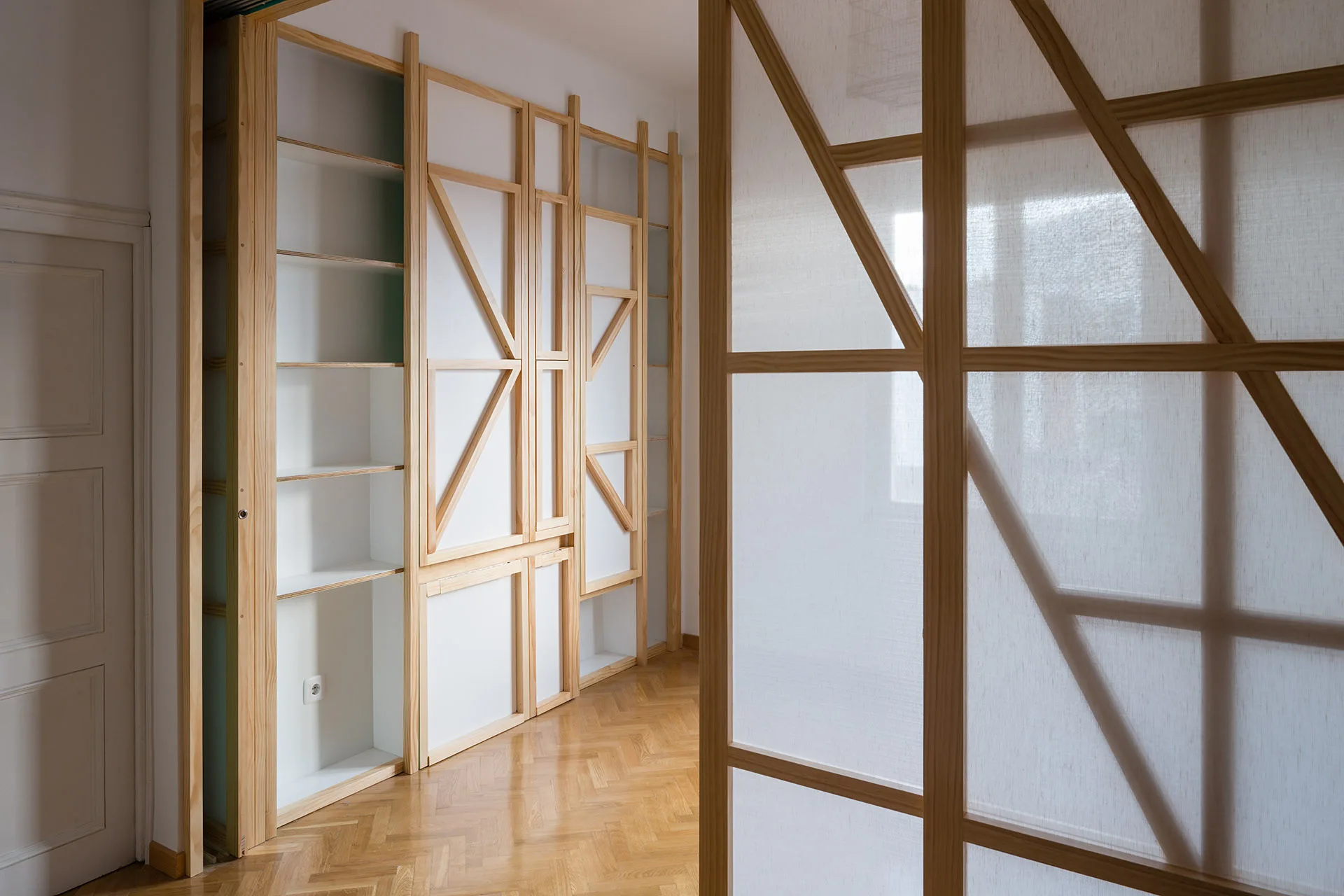 A minimalist room featuring custom light wood shelving units and a translucent screen room divider on herringbone patterned wood flooring.