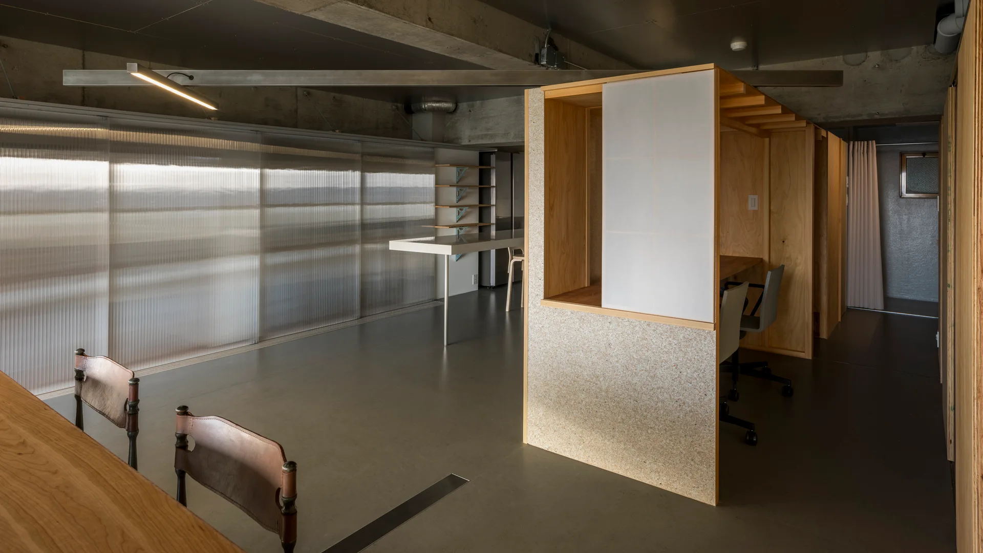 An industrial office interior features a custom plywood workstation partition, exposed concrete ceilings, and a ribbed translucent window screen.