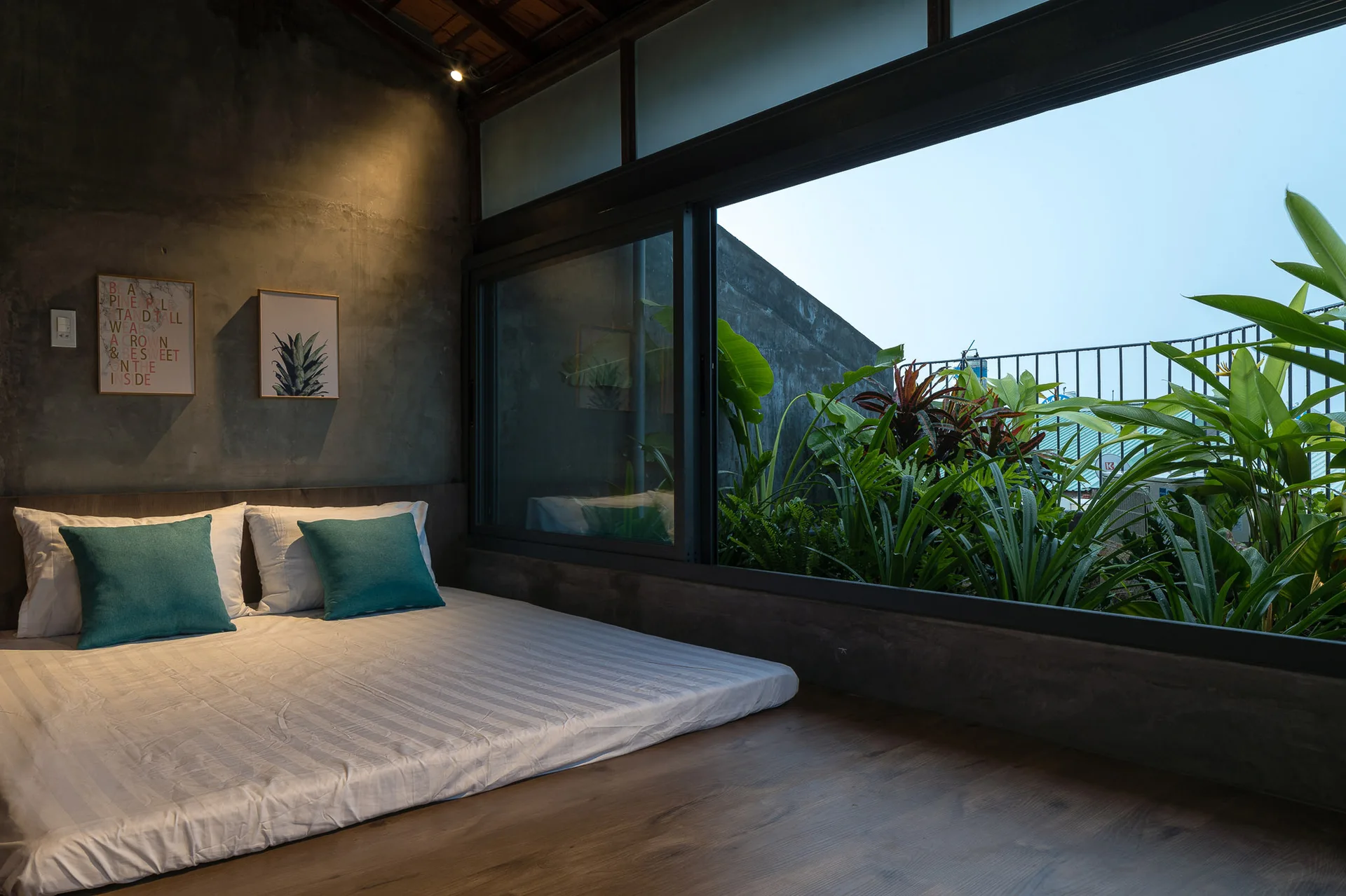A low-profile bed sits on a wood floor next to a dark concrete wall and a large window looking out at lush green tropical plants.