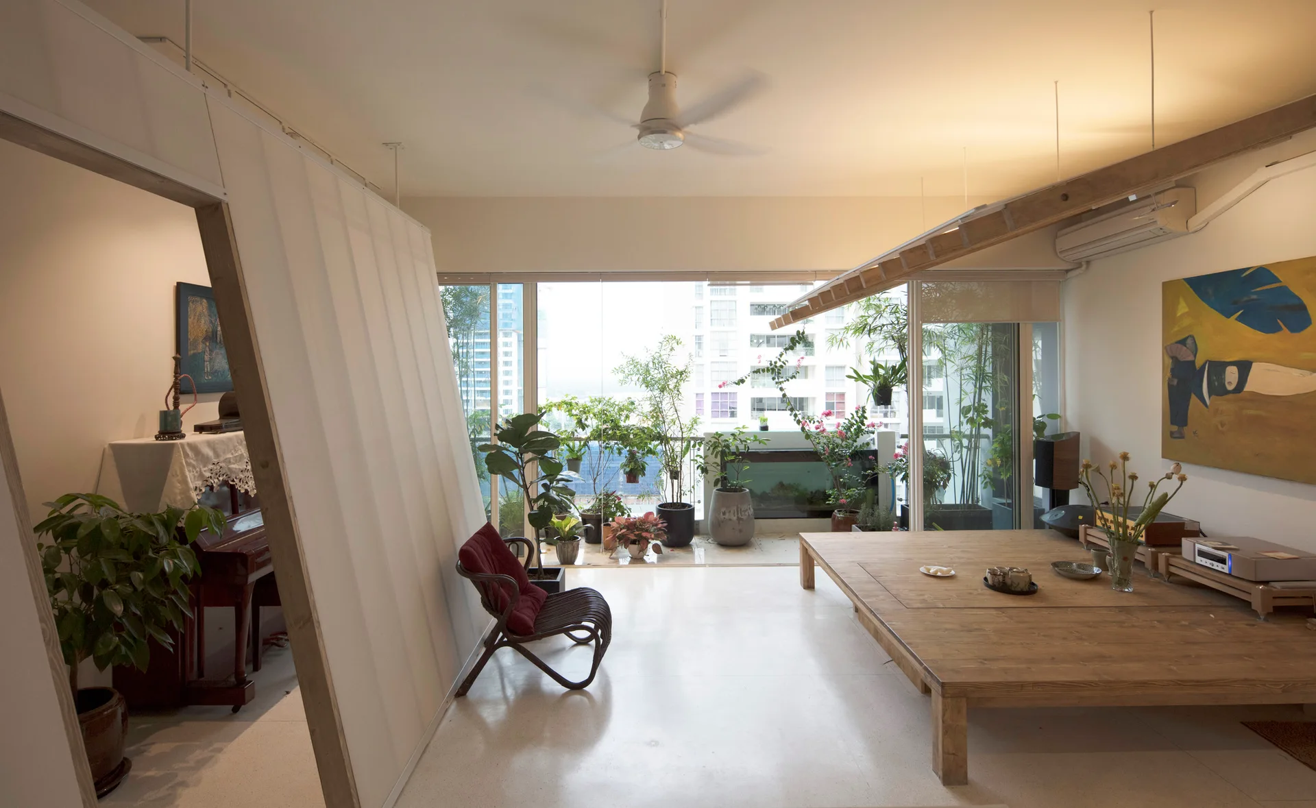 A minimalist living room features a large low wooden table, an angled white partition, and floor-to-ceiling windows opening to a green balcony.
