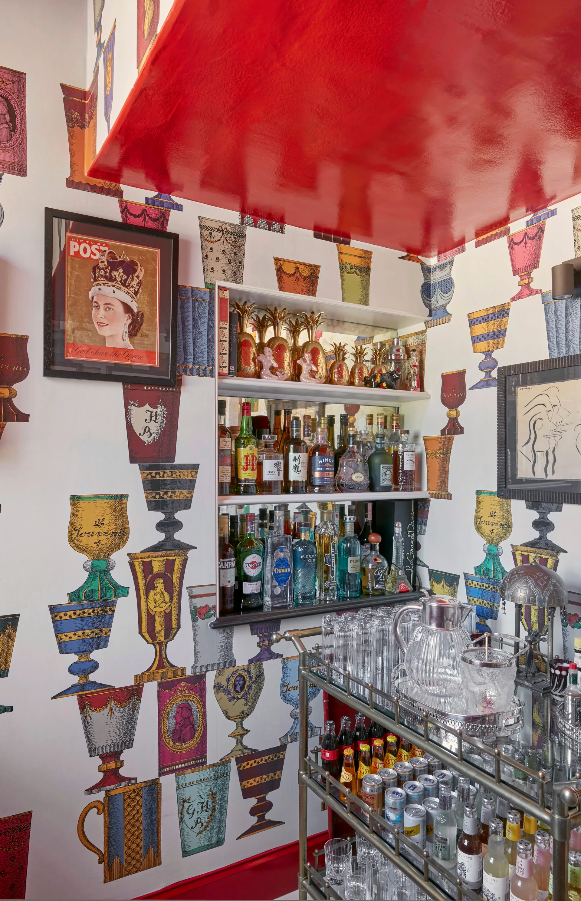 A home bar corner features goblet-patterned wallpaper, a high-gloss red ceiling, recessed wall shelving, and a metal bar cart stocked with bottles.